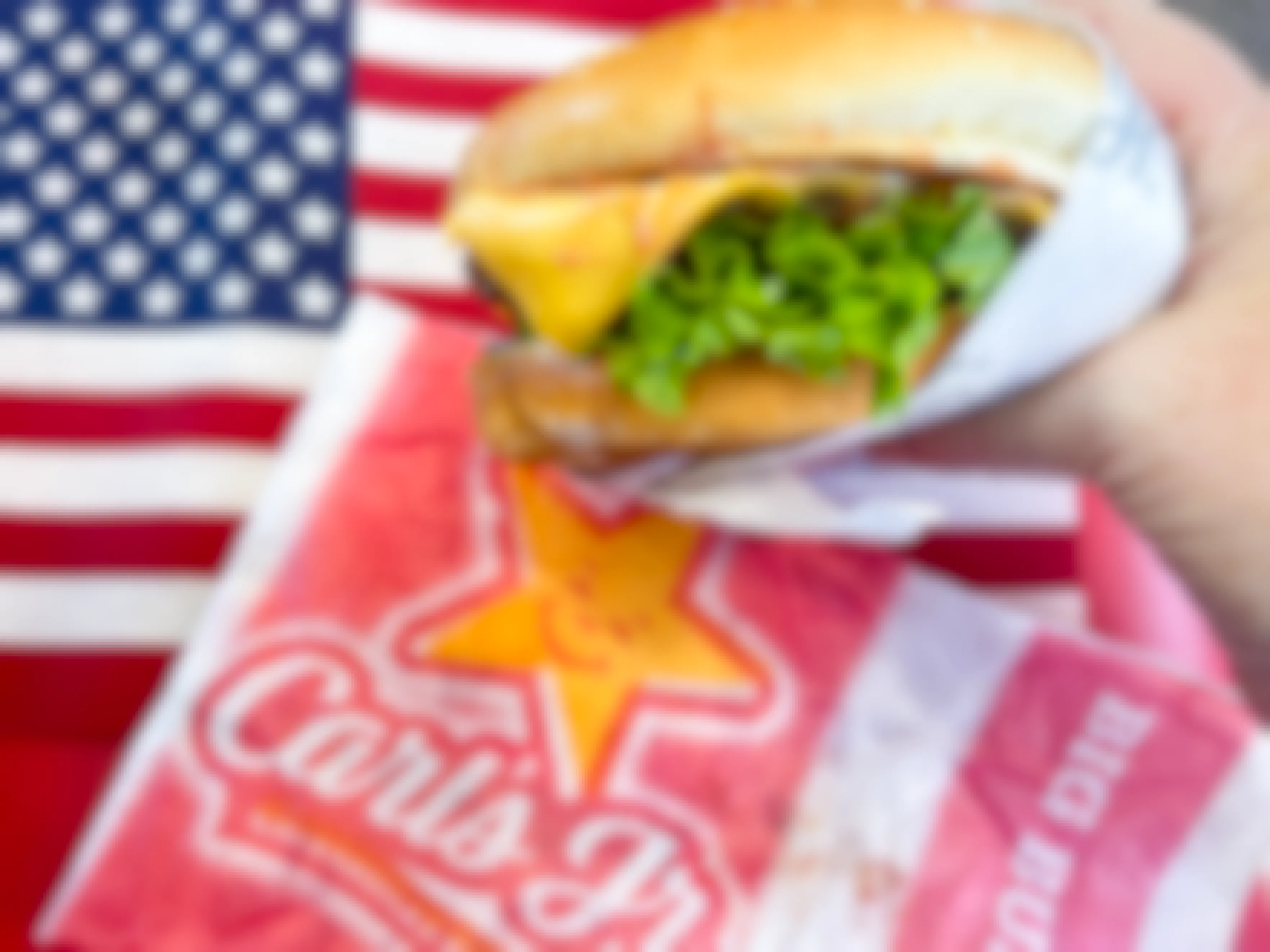 a hand holding a cheese burger in front of an american flag with a carls jr wrapper beside