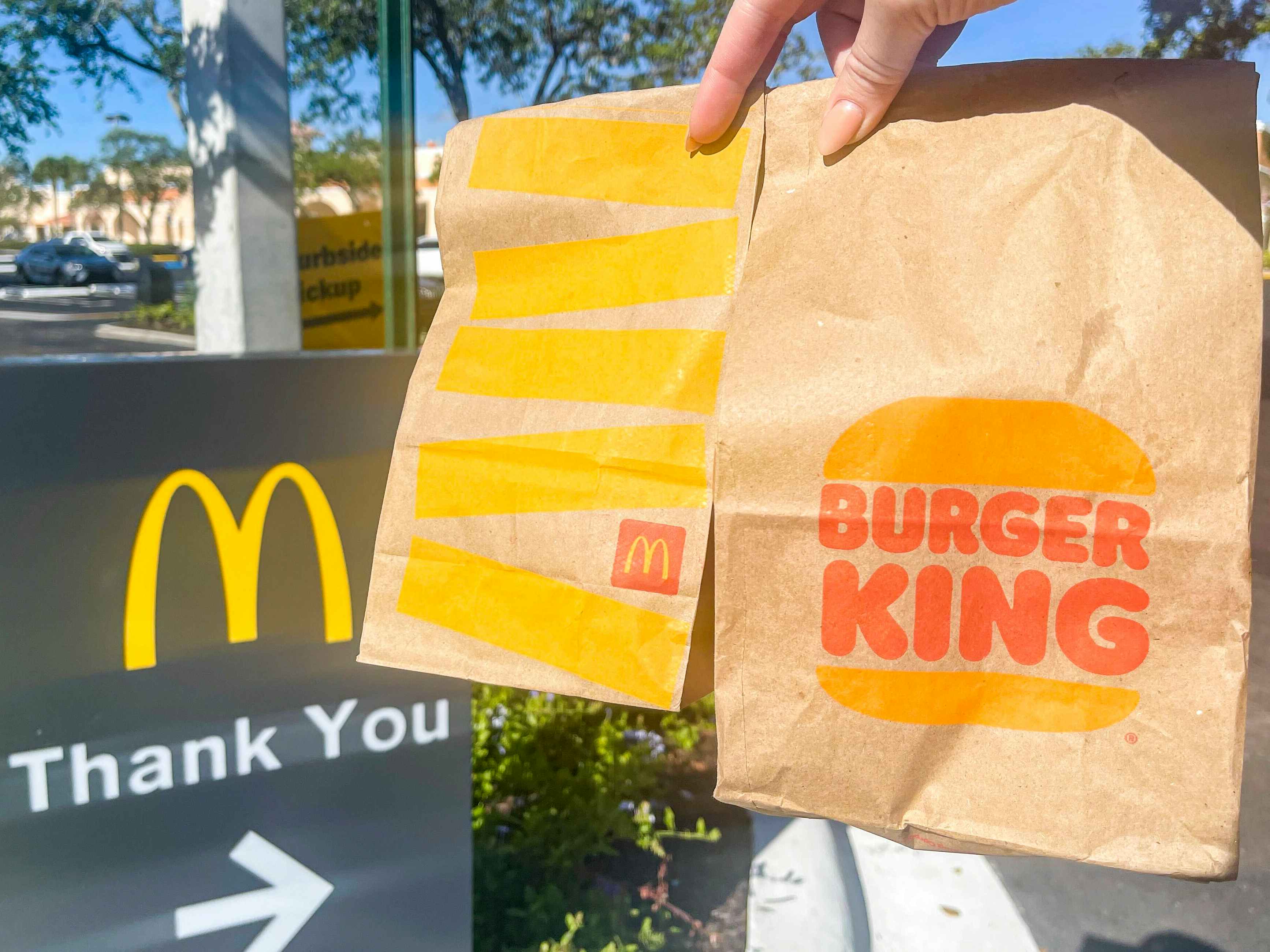 A person holding a McDonald's and Burger King to-go bag in one hand next to a sign that reads "thank you" with a McDonald's golden arch logo.