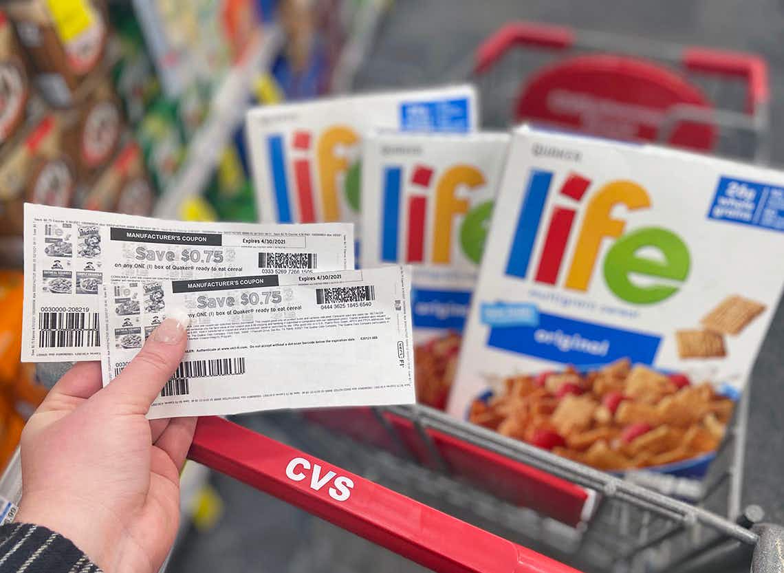 Hand holding two manufacturer coupons with three boxes of Life cereal in a CVS shopping cart