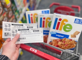 Hand holding two manufacturer coupons with three boxes of Life cereal in a CVS shopping cart