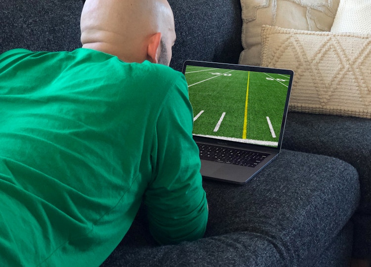 A man laying on a couch watching football on a laptop.