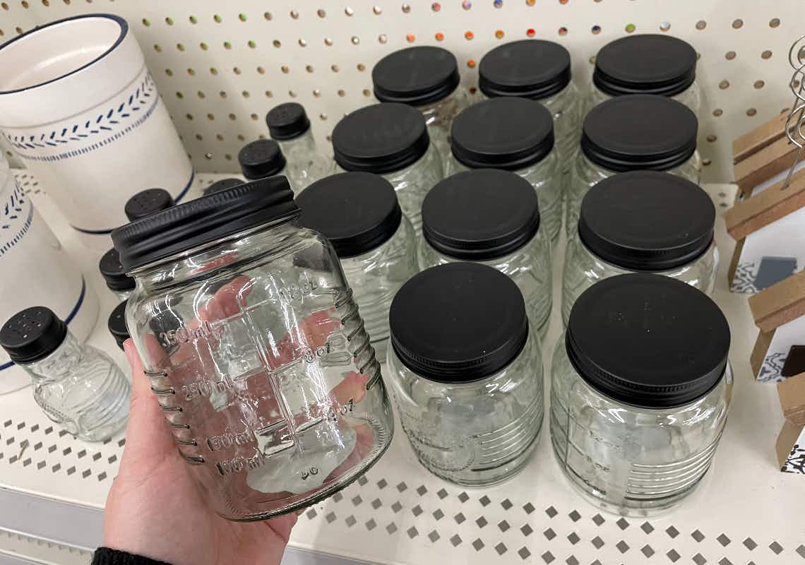 A person holding a glass jar with a black lid in front of a shelf with more glass jars on it.