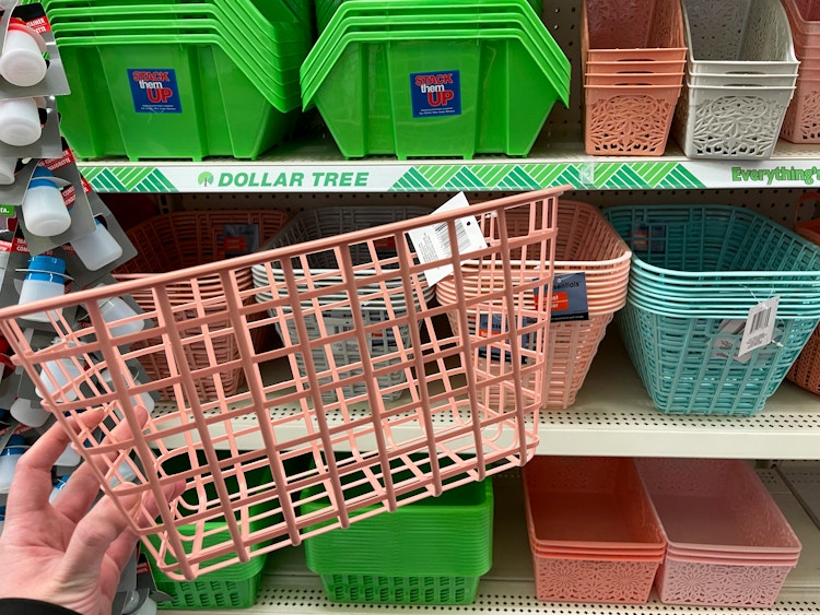 A person holding a pink storage basket in front of a dollar store shelf filled with other storage baskets.