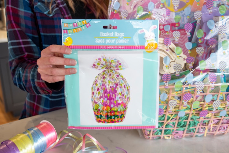 A woman holds up a package that says basket bags, two pack, in front of an Easter basket and a spool of pastel colored ribbon.