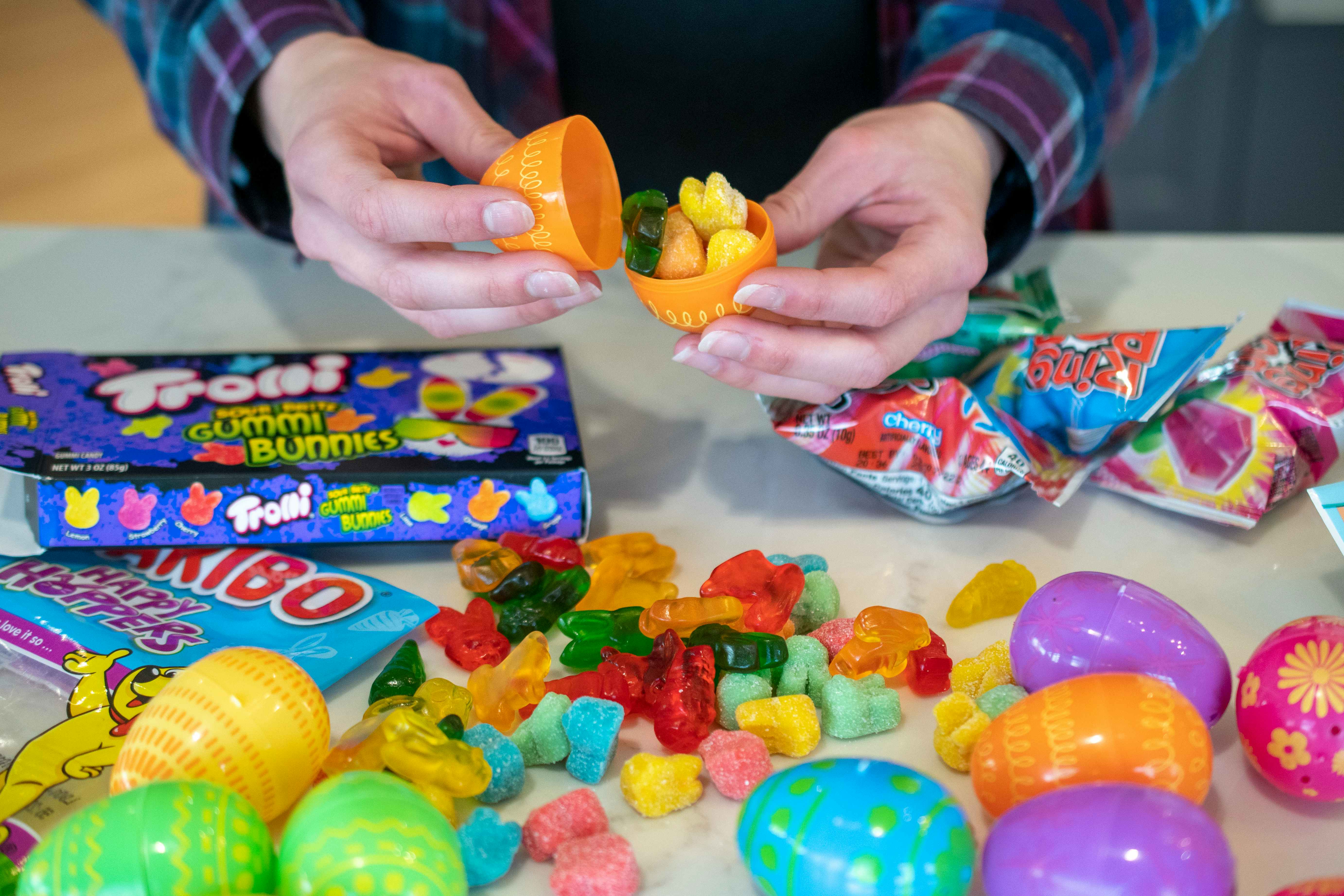 A pile of candy is on a countertop surrounded by plastic Easter eggs. A person is filling an egg with some of the candy.