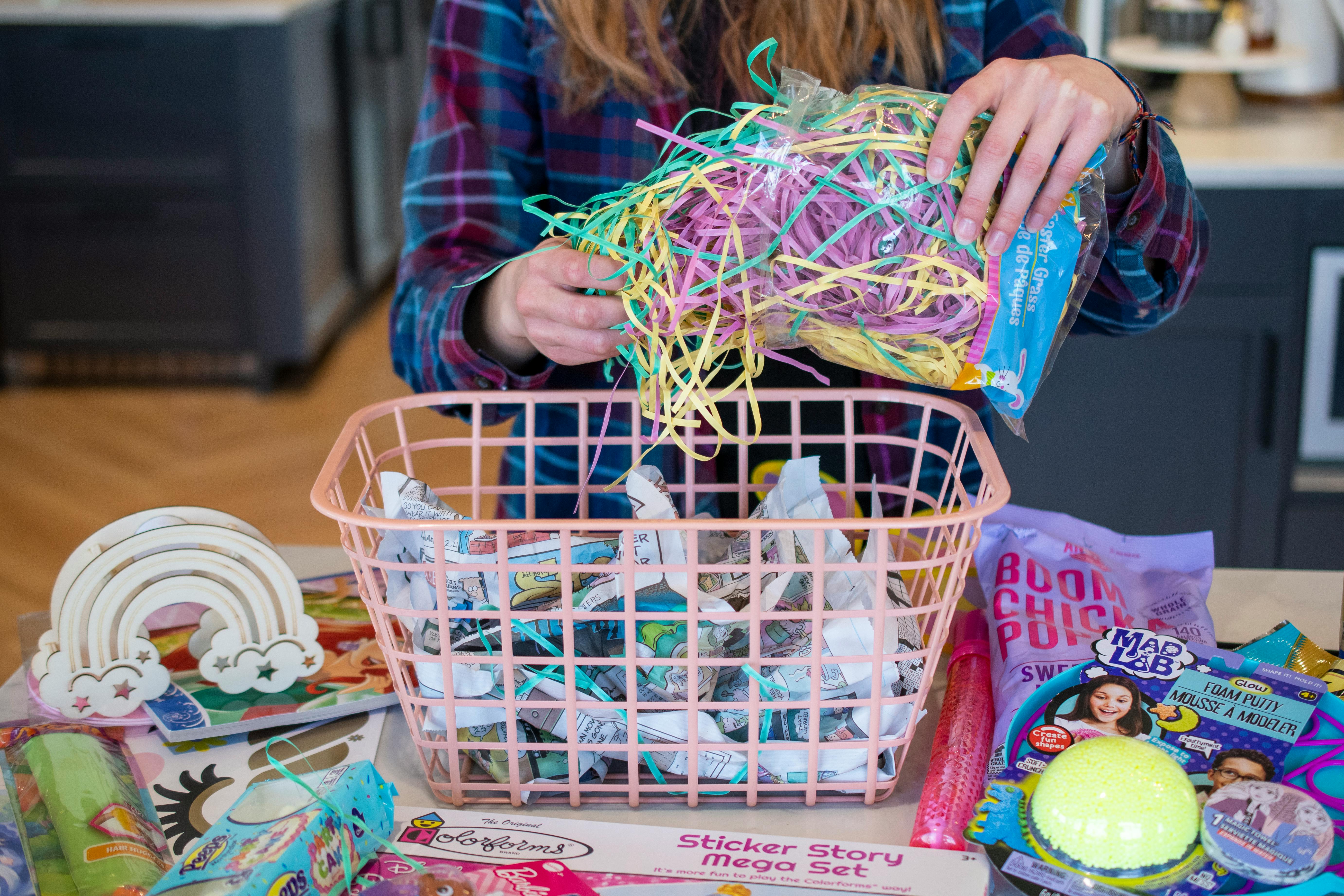 Toys and items are on the counter surrounding a pink basket. Newspaper is crumpled up inside of the basket and a person is pulling Easter grass from the packaging to put inside the basket.