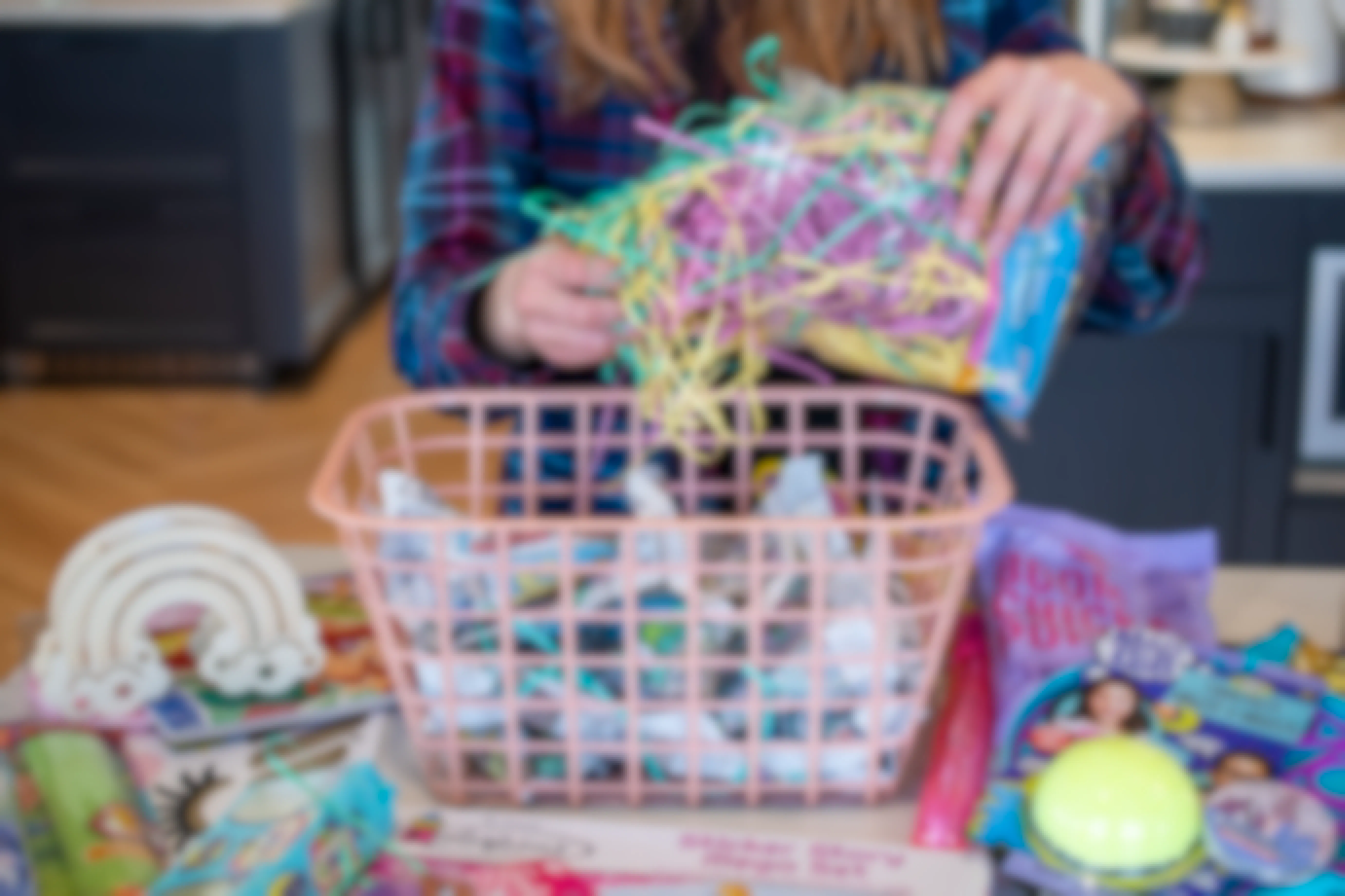 Toys and items are on the counter surrounding a pink basket. Newspaper is crumpled up inside of the basket and a person is pulling Easter grass from the packaging to put inside the basket.