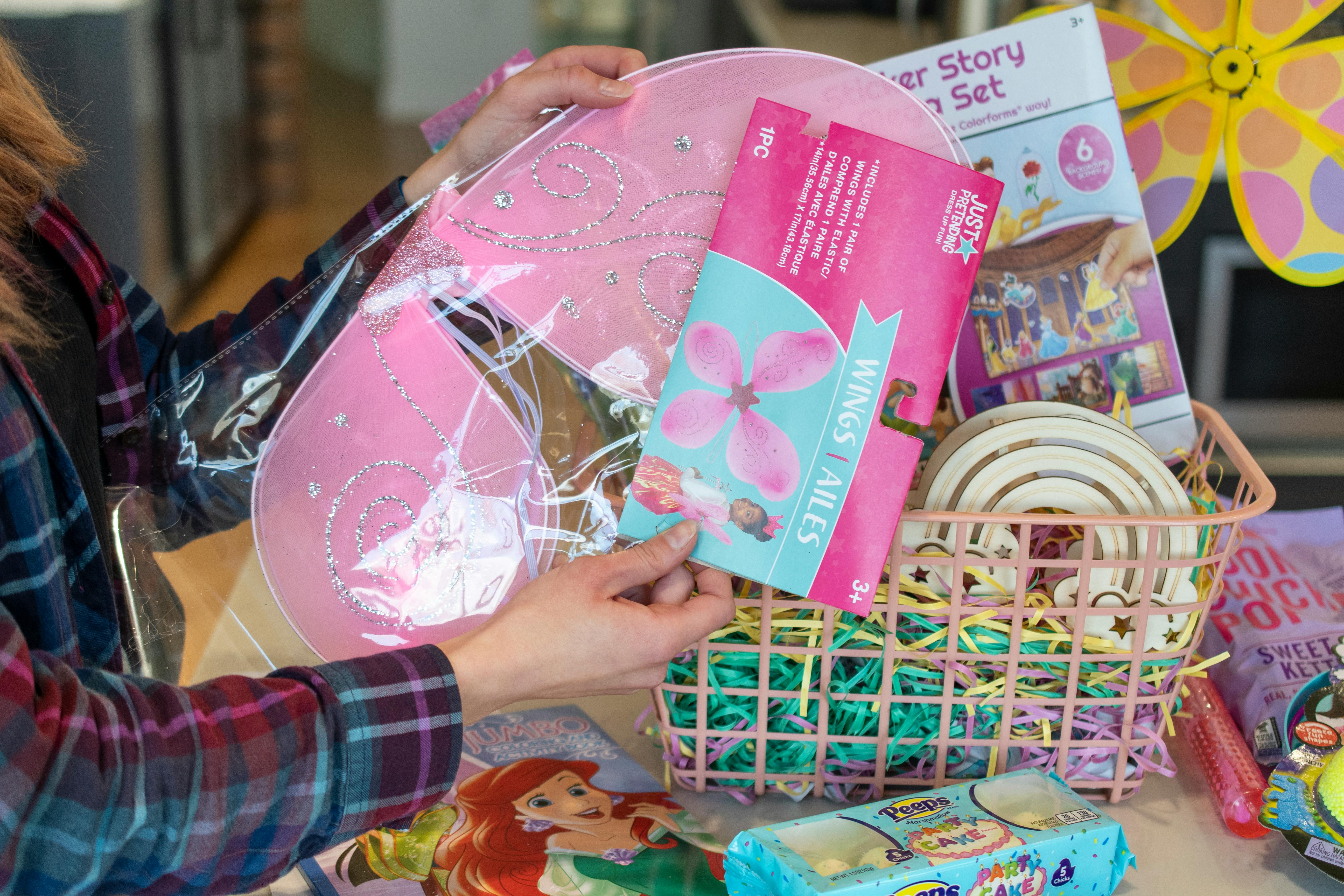 A person removing pink dress up wings from their packaging. An Easter basket filled with other items is in the background.