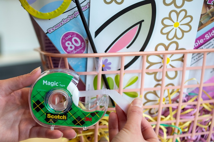 A person is pulling a piece of tape from the dispenser in front of a pink Easter basket filled with items.
