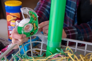 A person holding a roll of Scotch tape is using a piece to secure a green plastic Dollar Tree baseball bat to a gray basket.