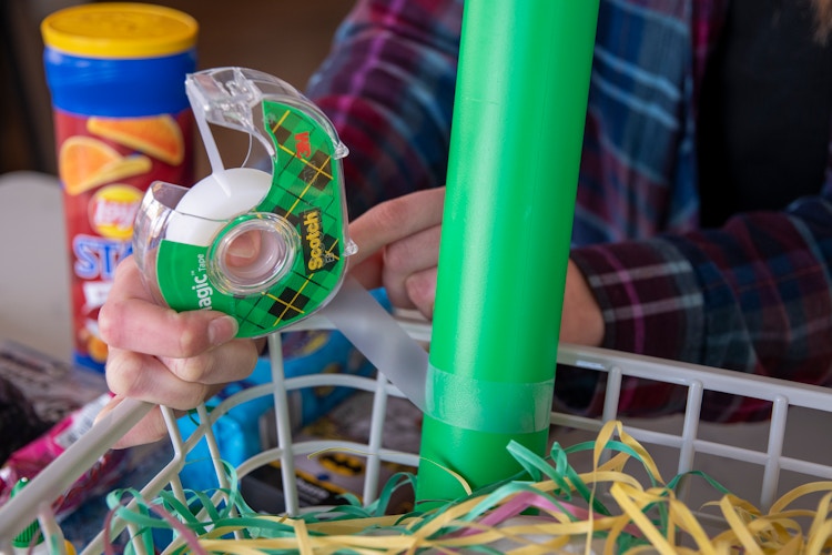 A person holding a roll of Scotch tape is using a piece to secure a green plastic Dollar Tree baseball bat to a gray basket.