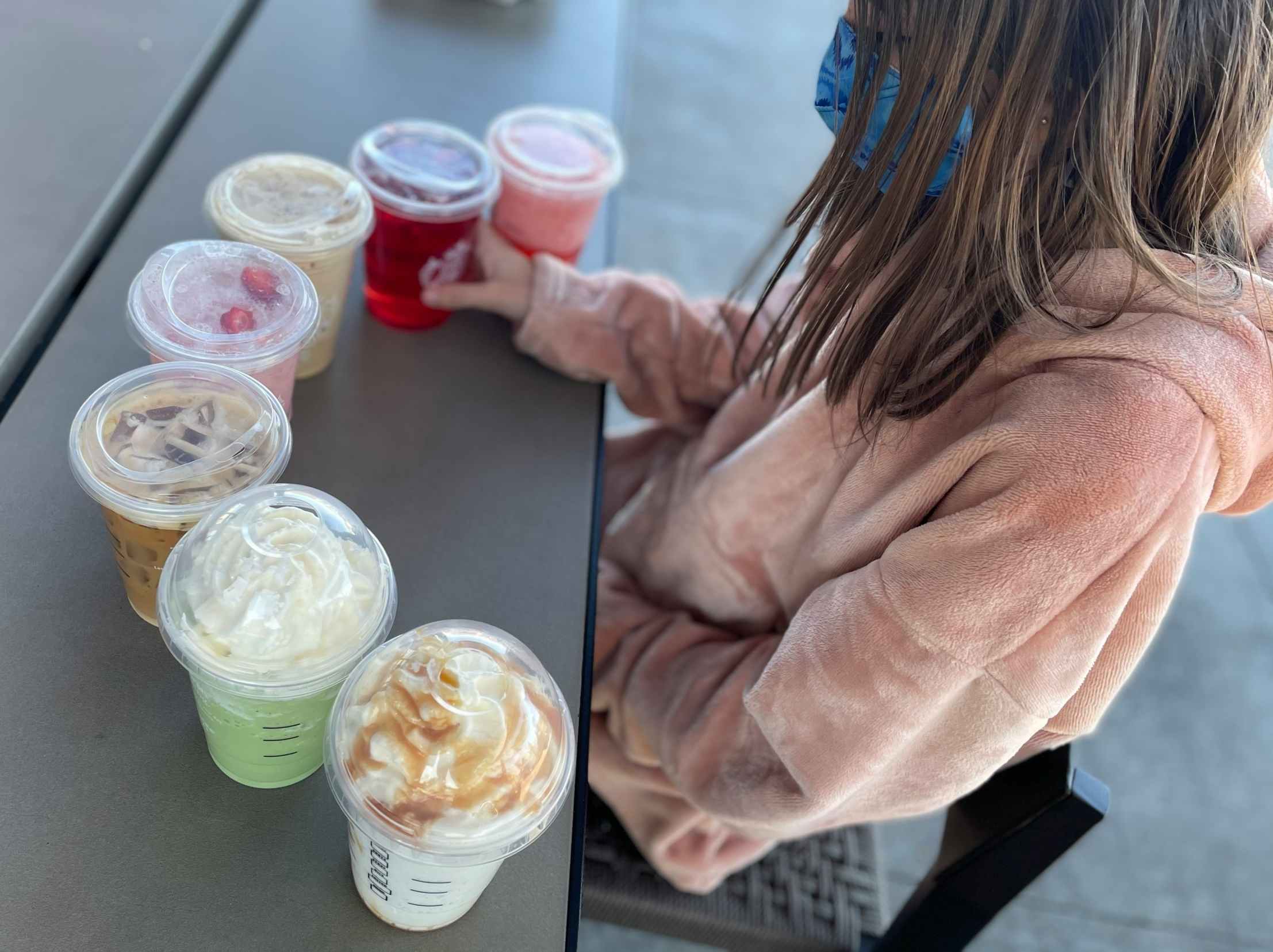 A young girl sitting at a table with a row of Starbucks secret menu drinks in front of her.