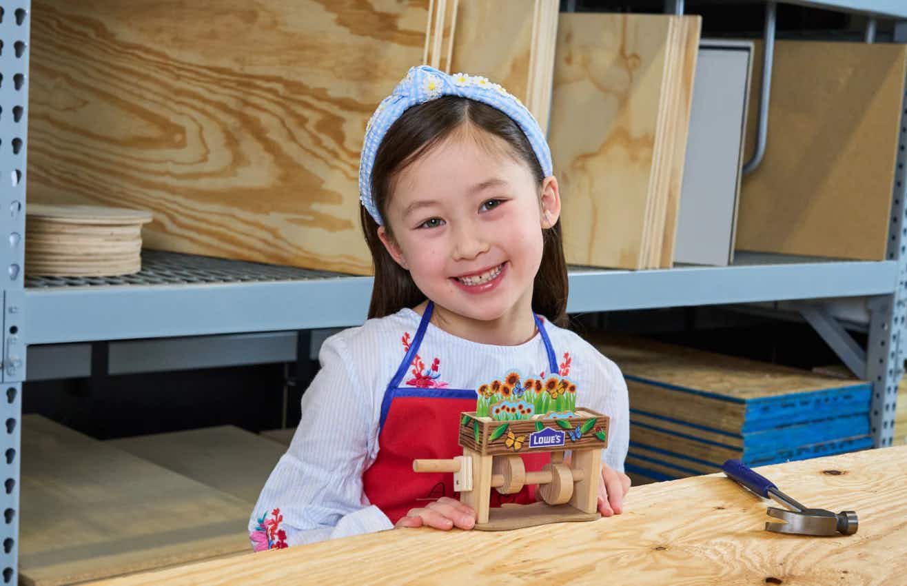 a young girl smiling and holding the DIY spring popup flower box she built in a Lowe's Kids' Workshop