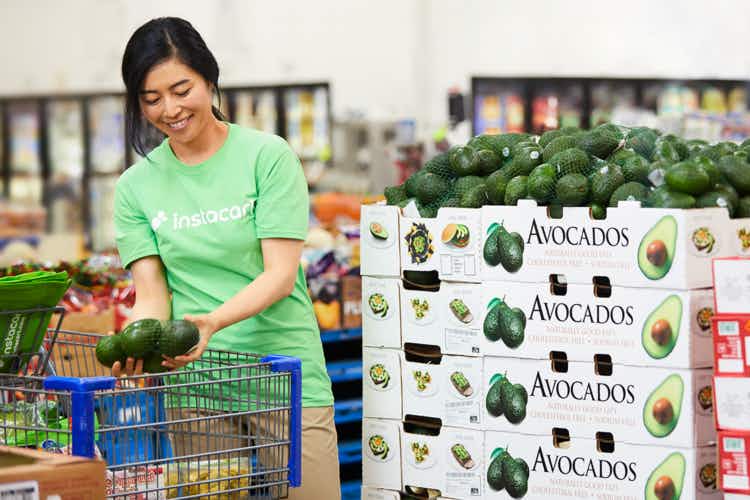 A woman in an Instacart shirt, putting avocados in a shopping cart inside Sam's Club