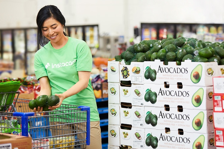 A woman in an Instacart shirt, putting avocados in a shopping cart inside Sam's Club