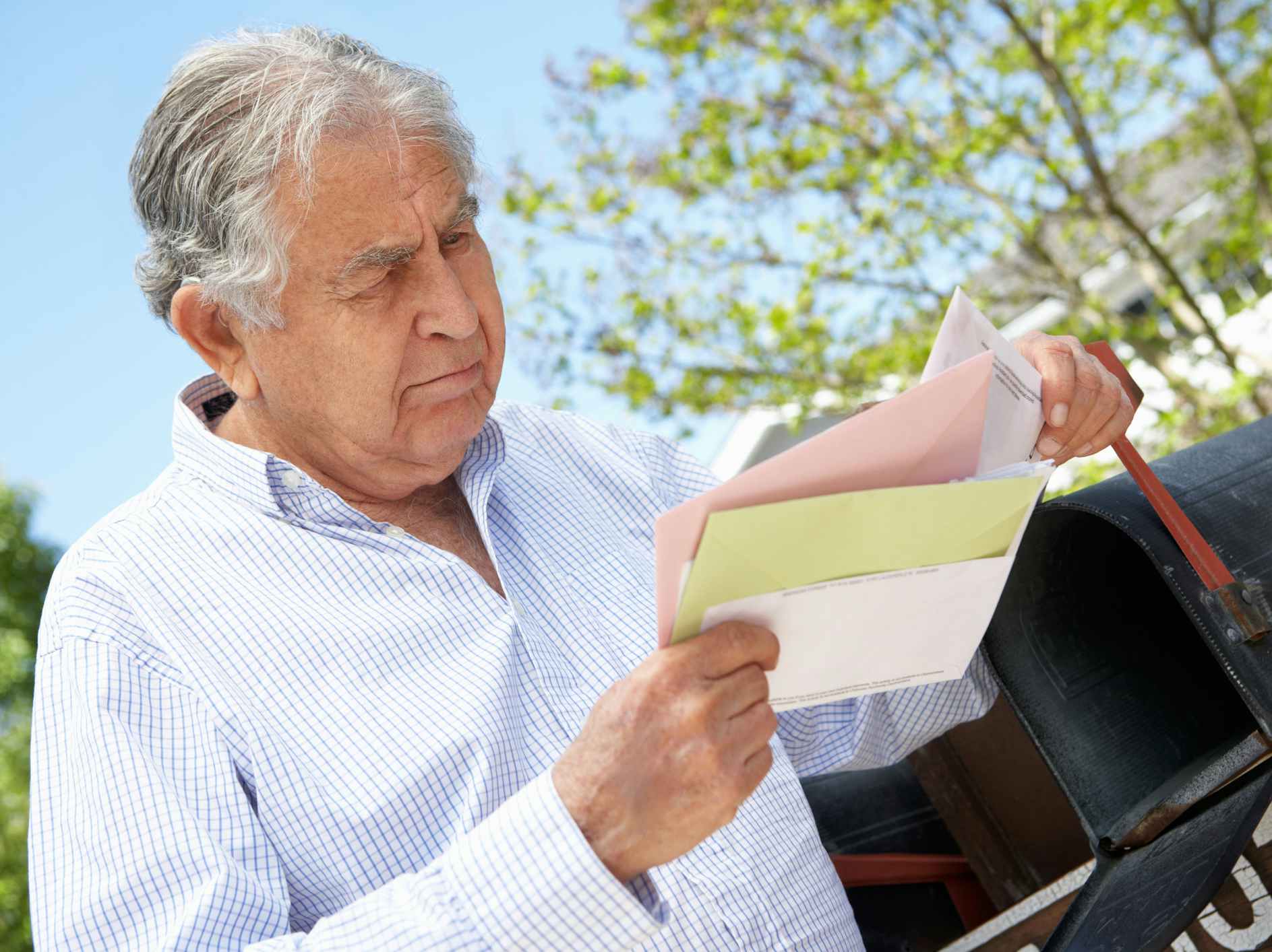 A senior man standing at a mailbox, looking at some envelopes.