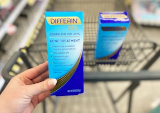A hand holding Differin gel in front of a shopping cart