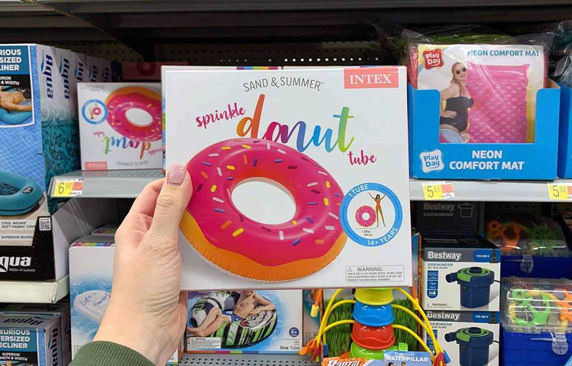 A person's hand holding up a boxed Intex Sand & Summer inflatable donut tube in front of a shelf in the seasonal section at Walmart.
