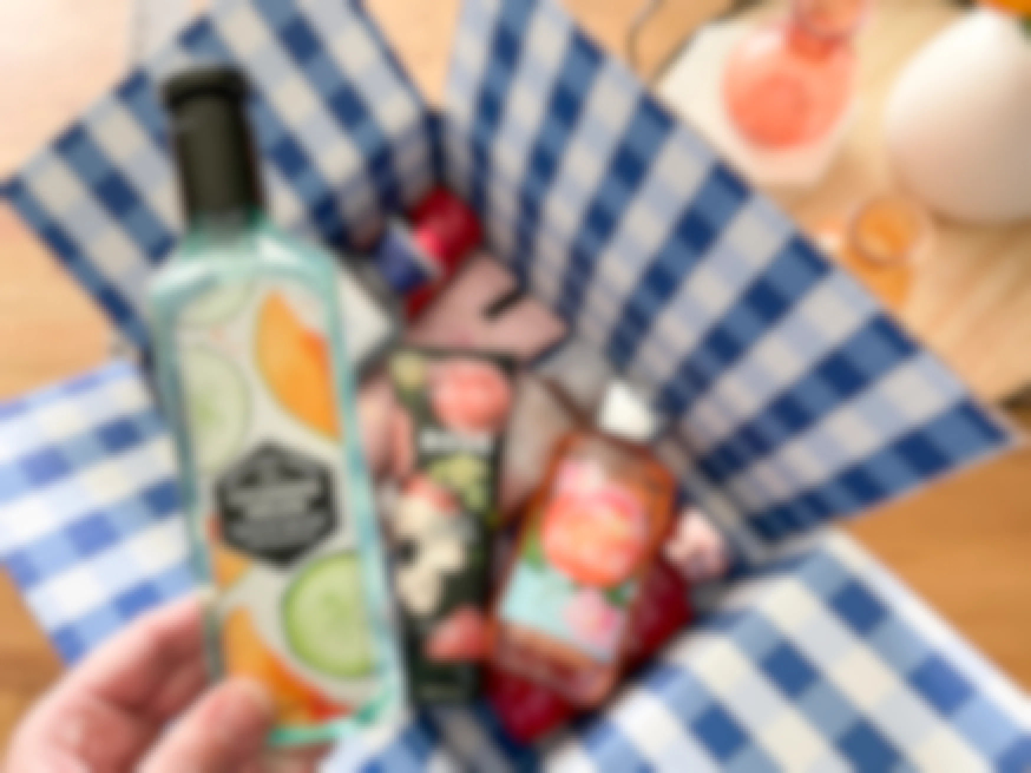 A Bath & body works online order box filled with products is open on a table. A person holds a bottle of Cucumber Melon hand soap next to the box, having just taken it out.