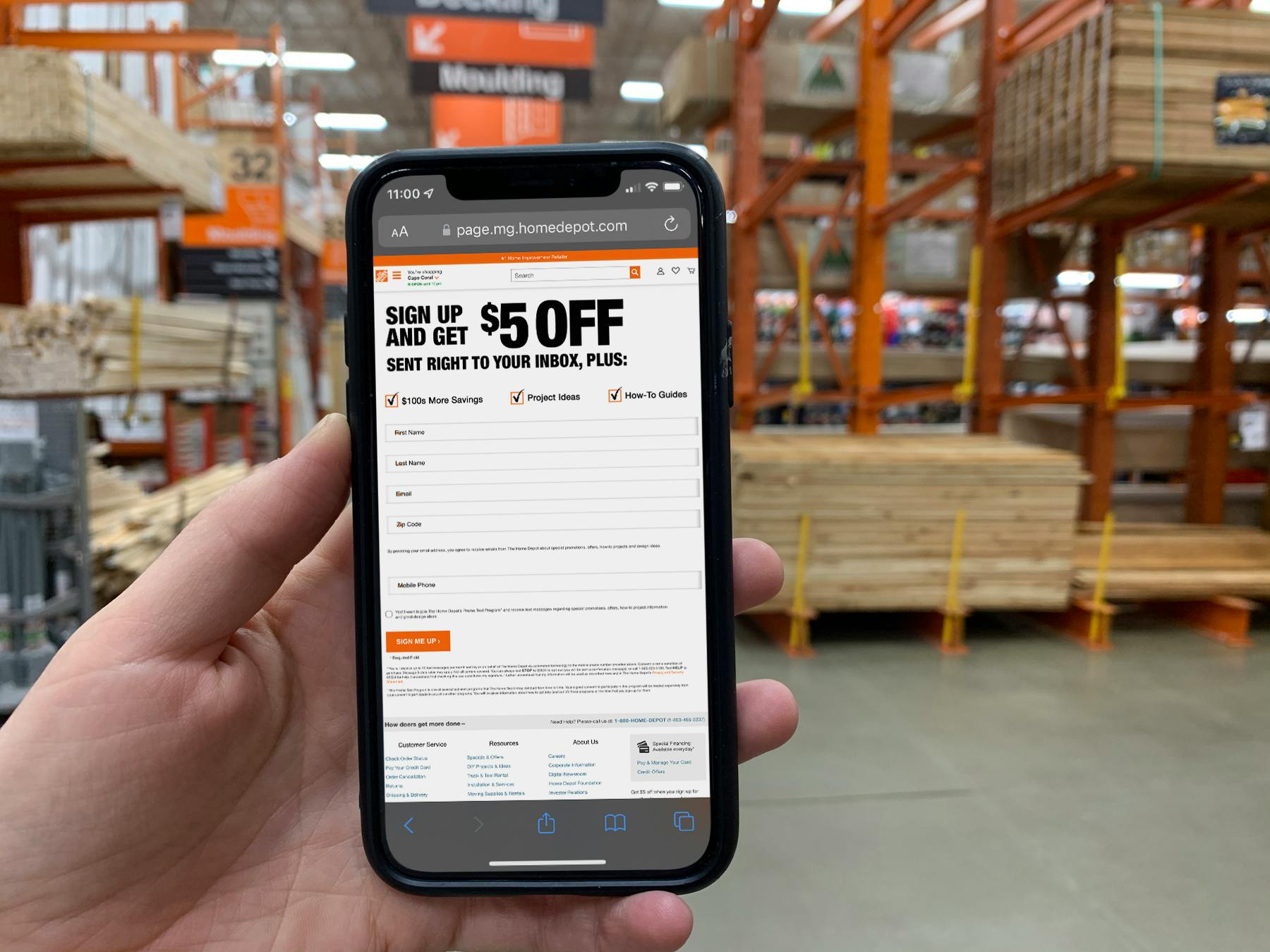 A person holding up a cell phone displaying the garden club sign up page on Home Depot's website offering a $5 off coupon
