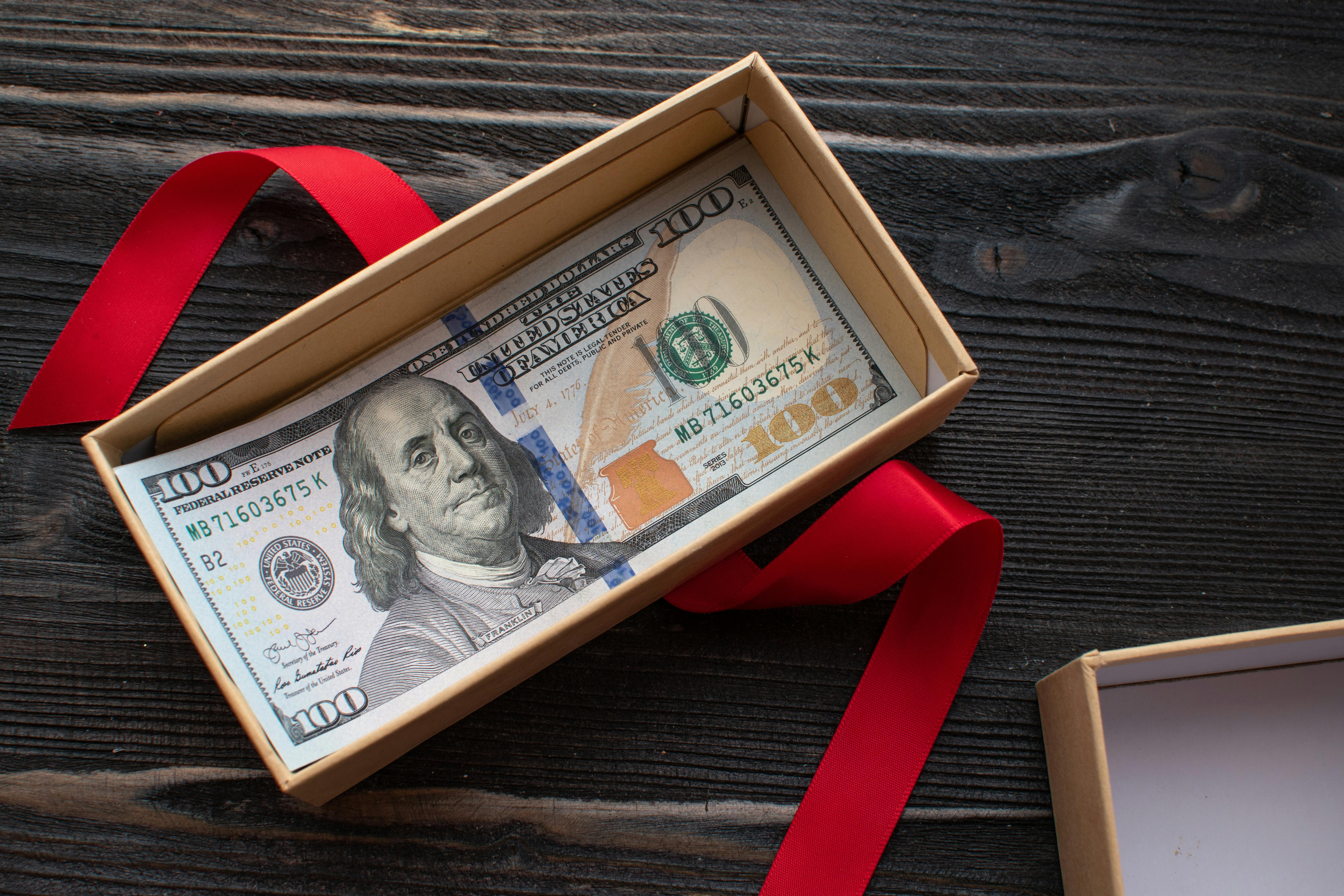 An opened gift box with 100 dollar bills in it, sitting on a wooden table.