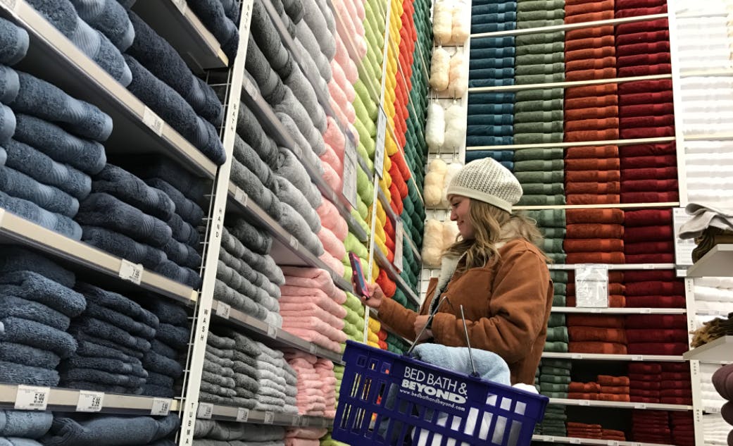A person holding a Bed Bath & Beyond shopping basket and looking at her phone while standing in the towel section of Bed Bath & Beyond with towels stocked on walls of shelves.