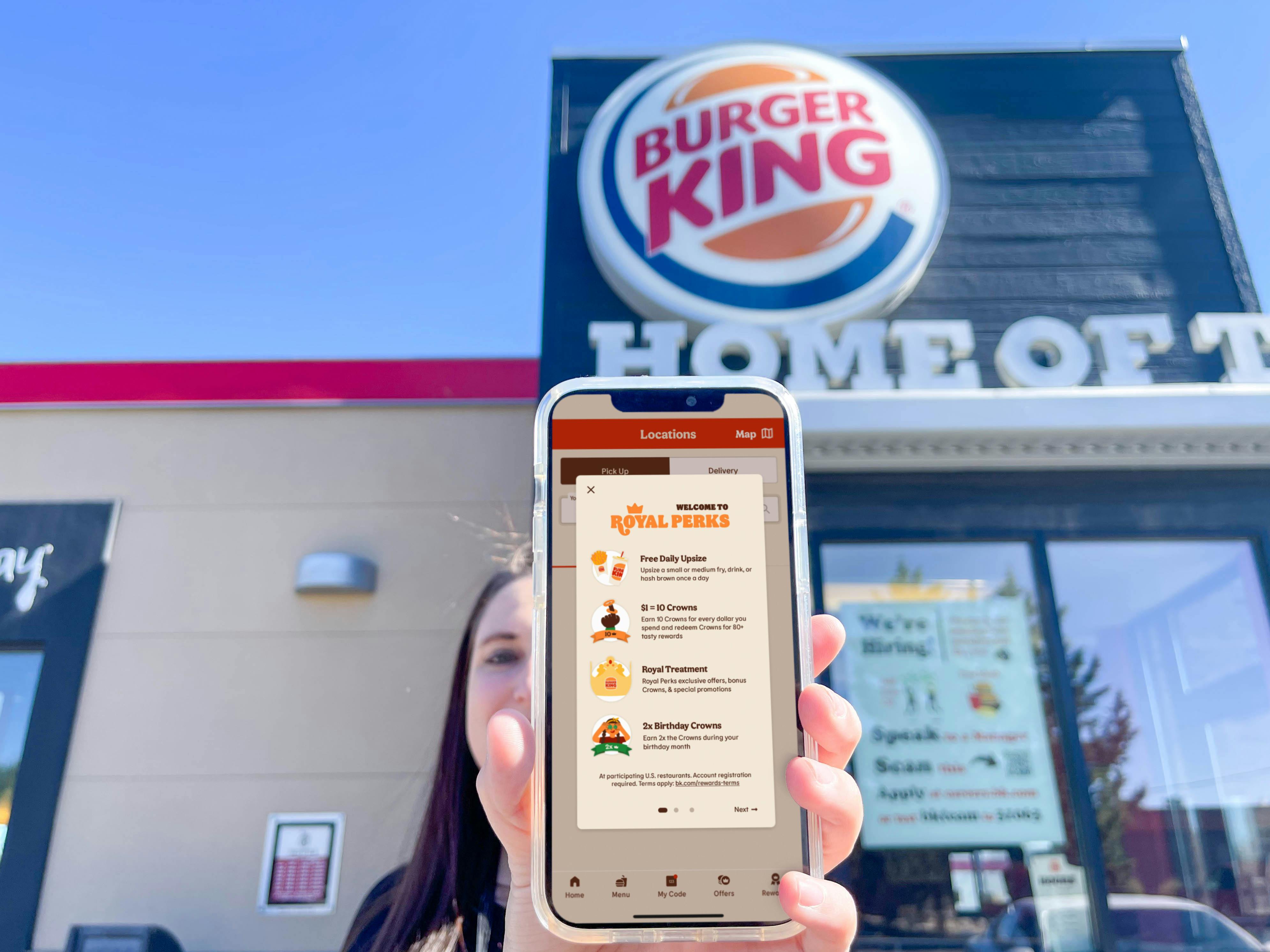 a woman holding a cellphone in front of face with burger king app on screen standing in front of burger king