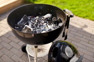 There is a charcoal grill with the top off standing on a bricked area outdoors next to the grass.
