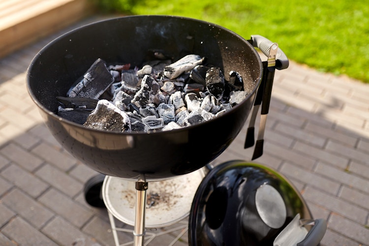 There is a charcoal grill with the top off standing on a bricked area outdoors next to the grass.