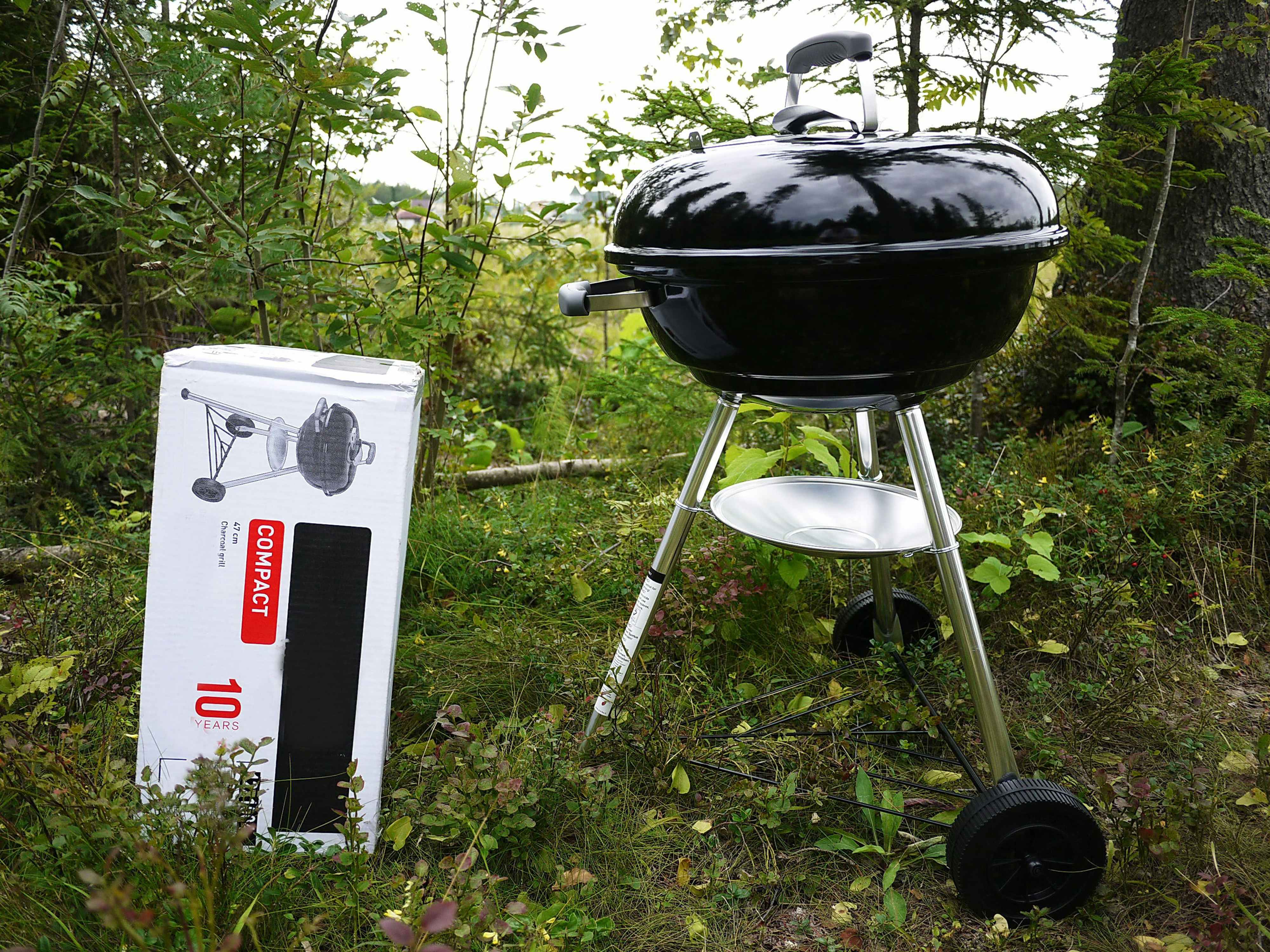Black grill next to a box that says compact with a picture of the grill on it. The grill and box are outside surrounded by grass and trees.