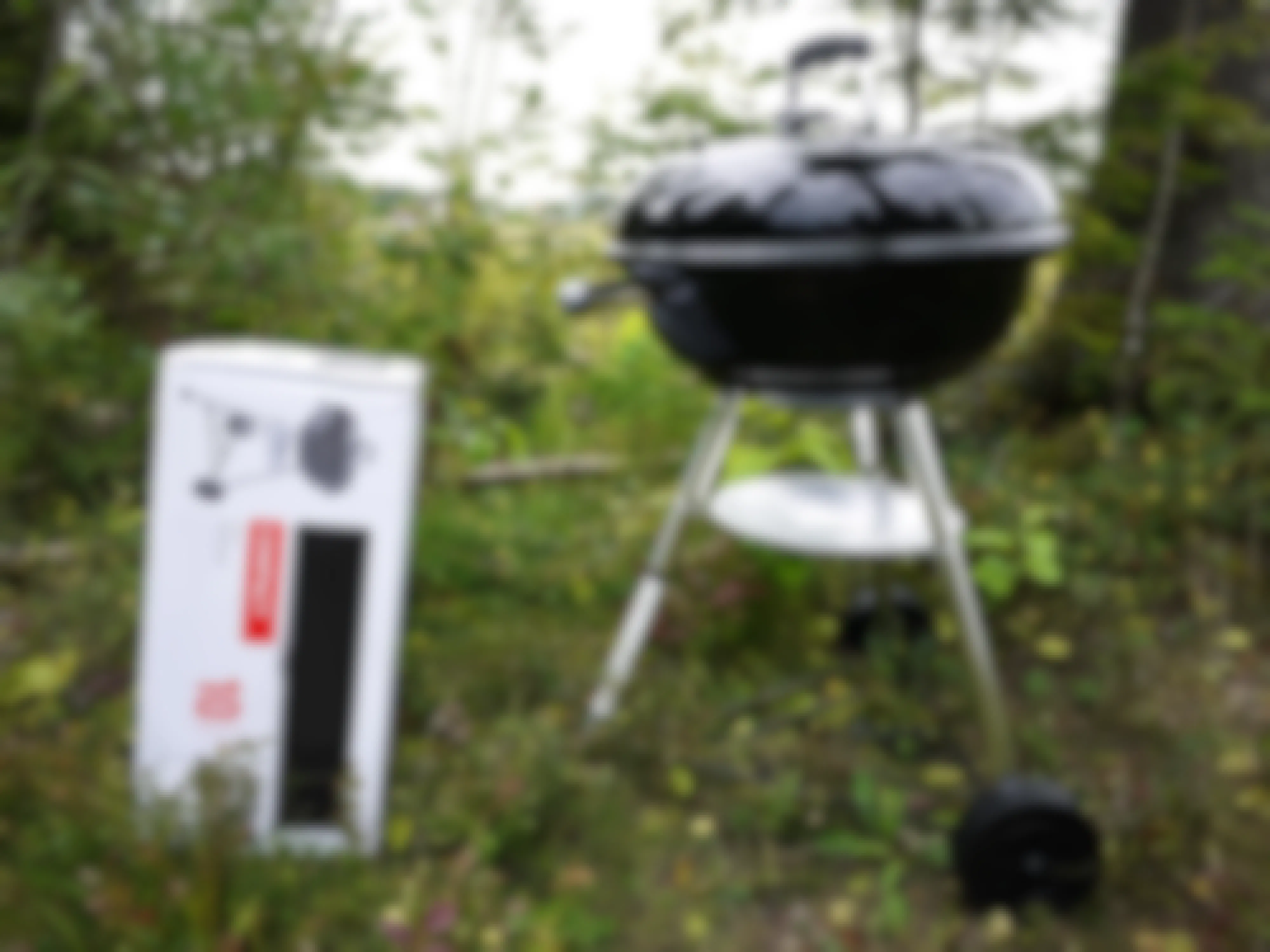 Black grill next to a box that says compact with a picture of the grill on it. The grill and box are outside surrounded by grass and trees.