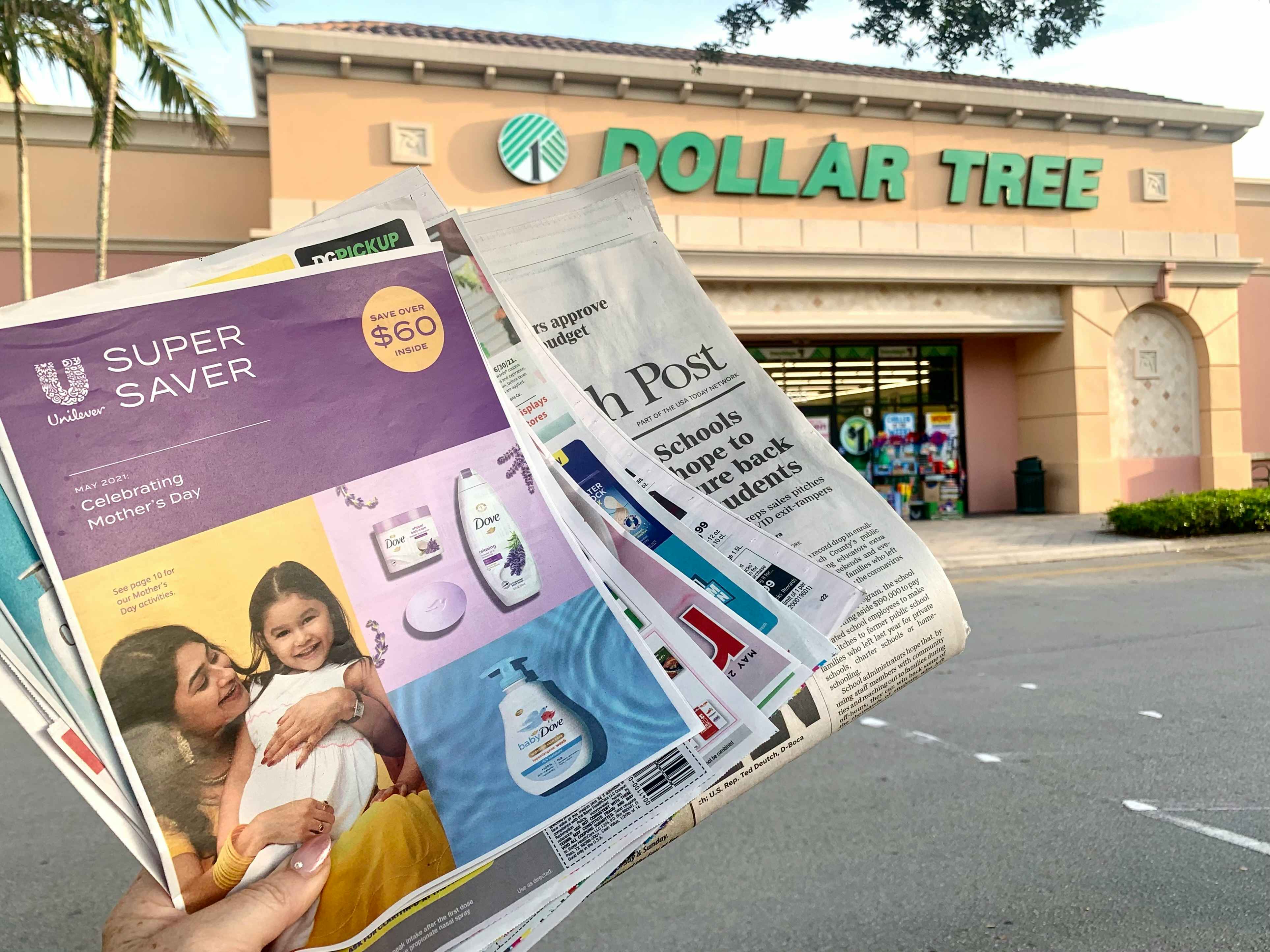 A person's hand holding a stack of coupon leaflets and a newspaper in front of a Dollar Tree storefront.