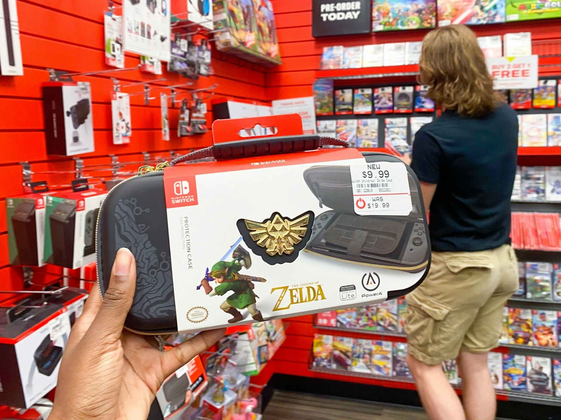 A person's hand holding up a Nintendo Switch Legend of Zelda carrying case inside a GameStop store with someone looking at games in the background.