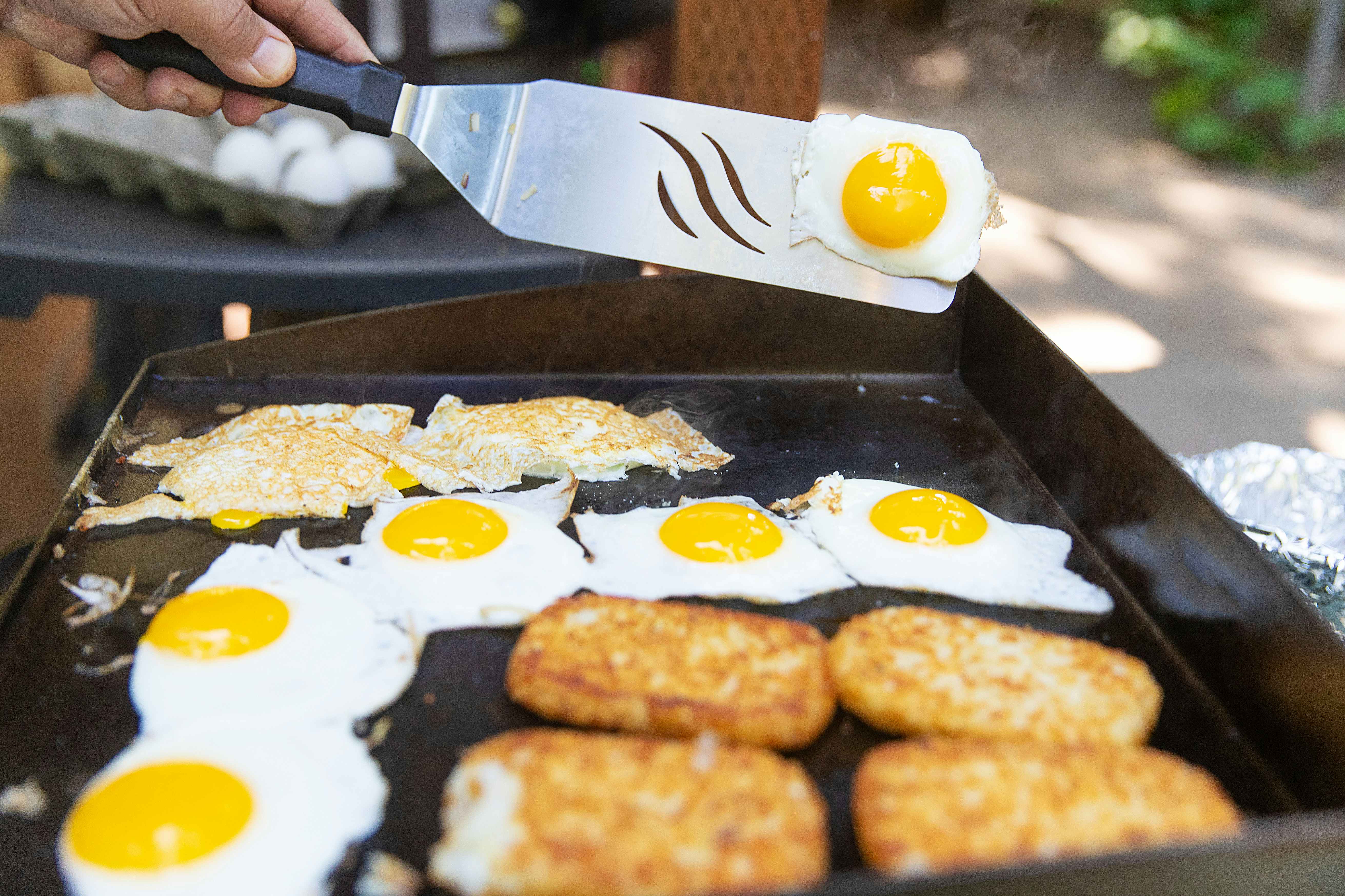 Eggs and hashbrowns are being cooked on a griddle. Someone is holding a spatula and flipping an egg.