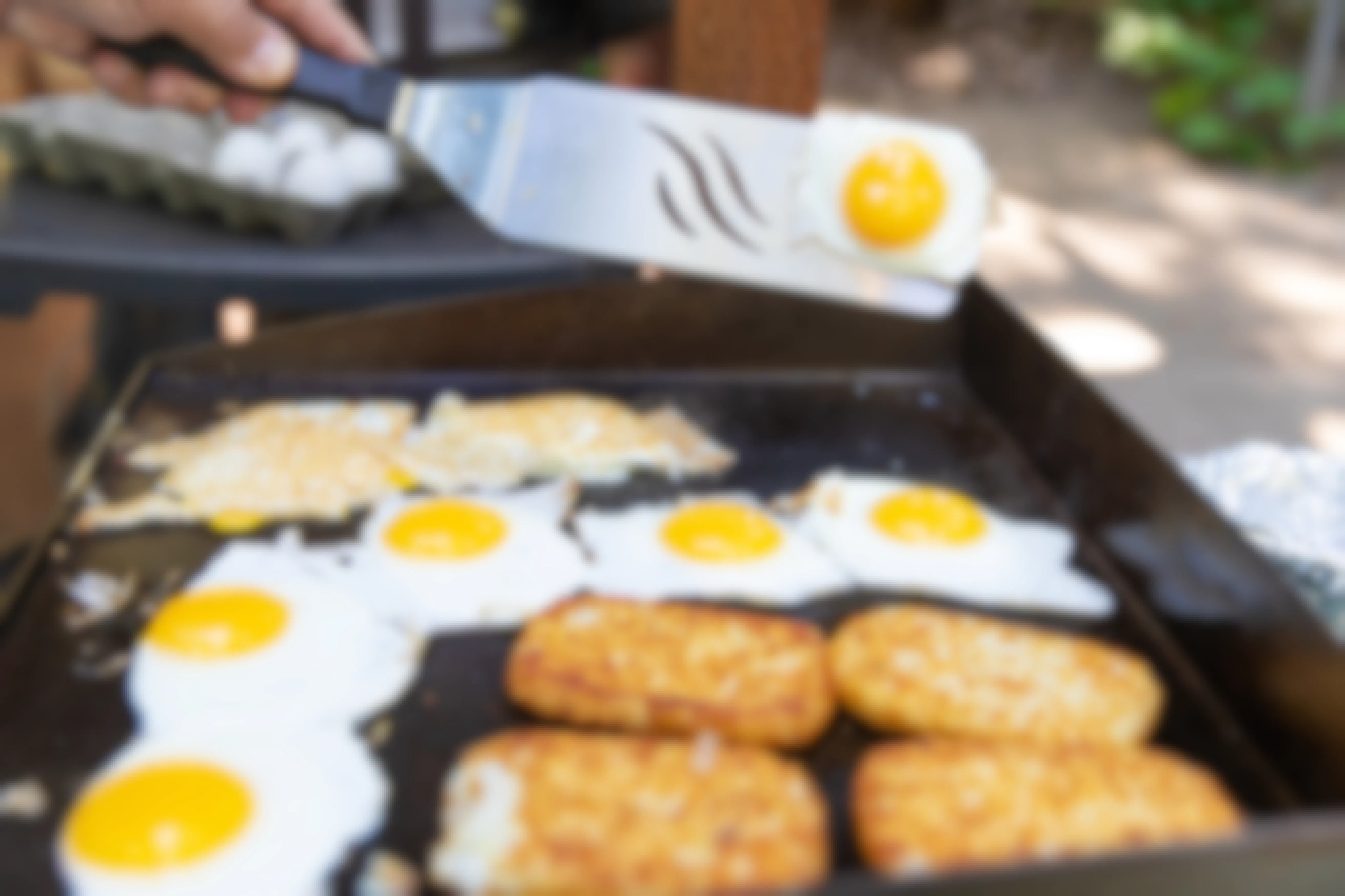 Eggs and hashbrowns are being cooked on a griddle. Someone is holding a spatula and flipping an egg.