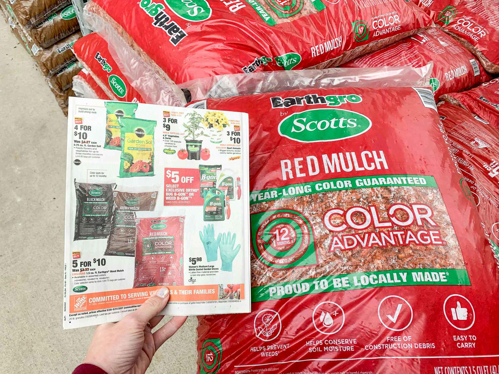 A hand holding a home Depot Memorial Day Sale ad next to a bag of Earthgro by Scotts red mulch.
