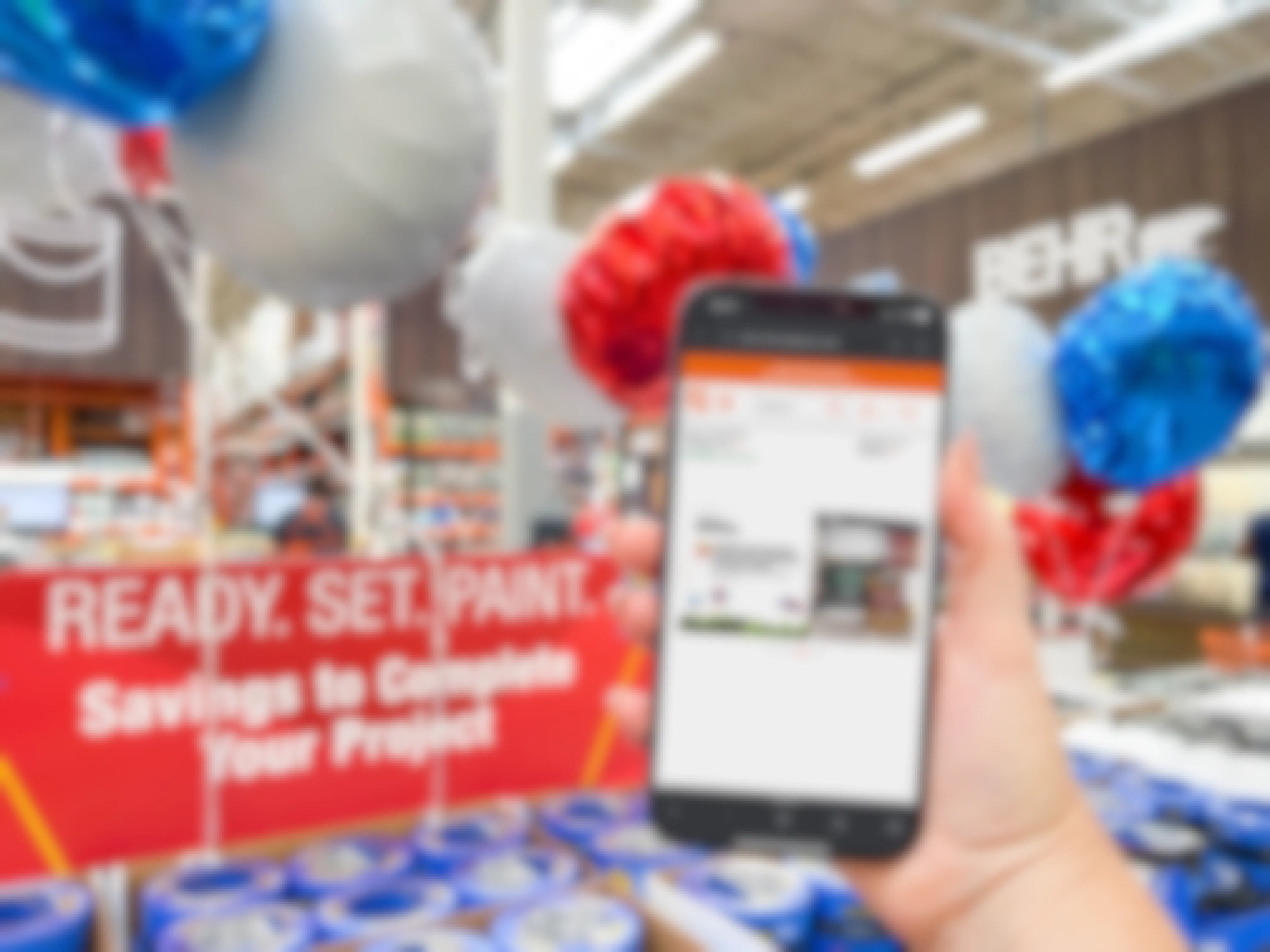 A person's hand holding up an iPhone displaying the Home Depot memorial day paint sale in the Home Depot app in front of a paint display with red, white, and blue balloons.