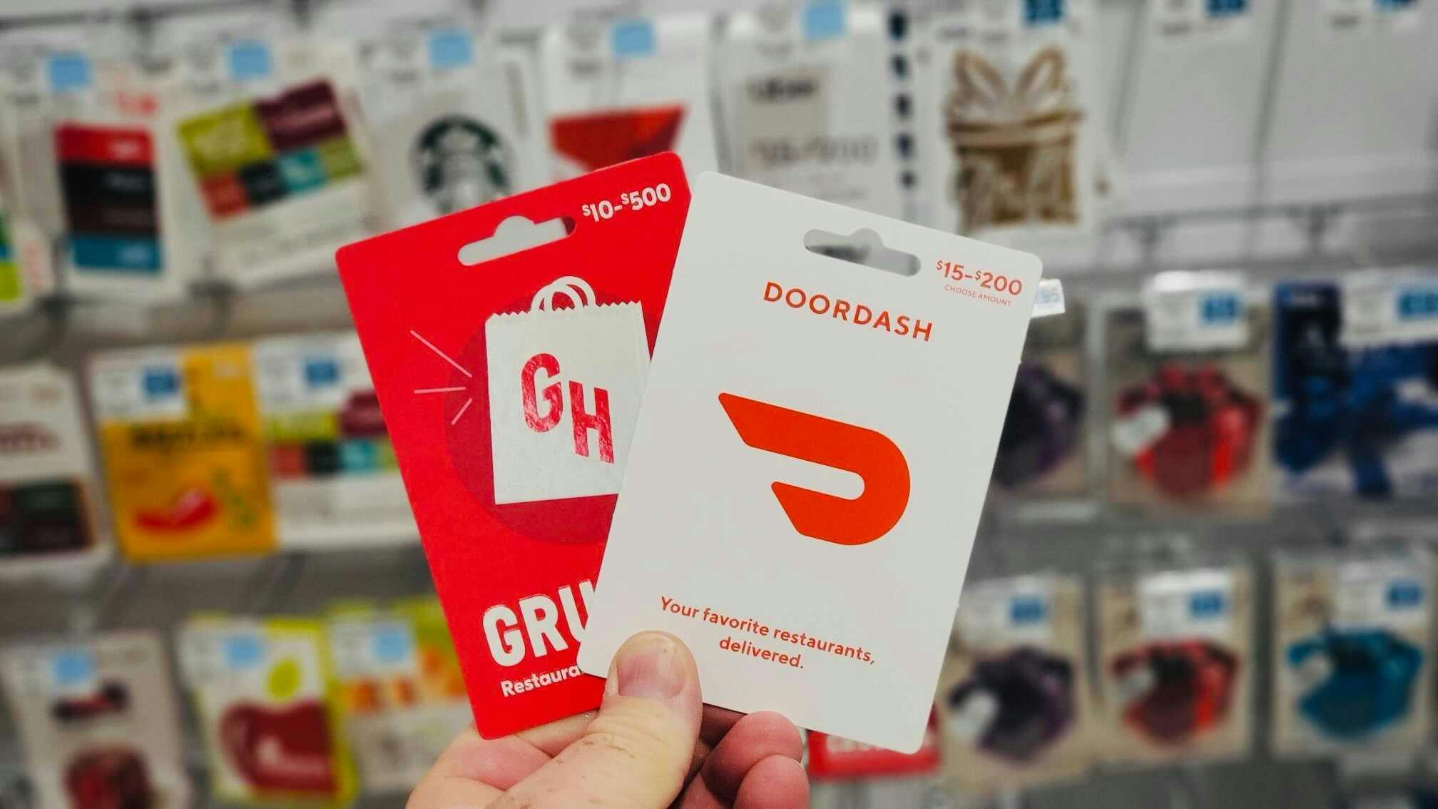 - Two gift cards being held center-frame in front of a wall of gift cards, one is for Grubhub and one for Doordash.