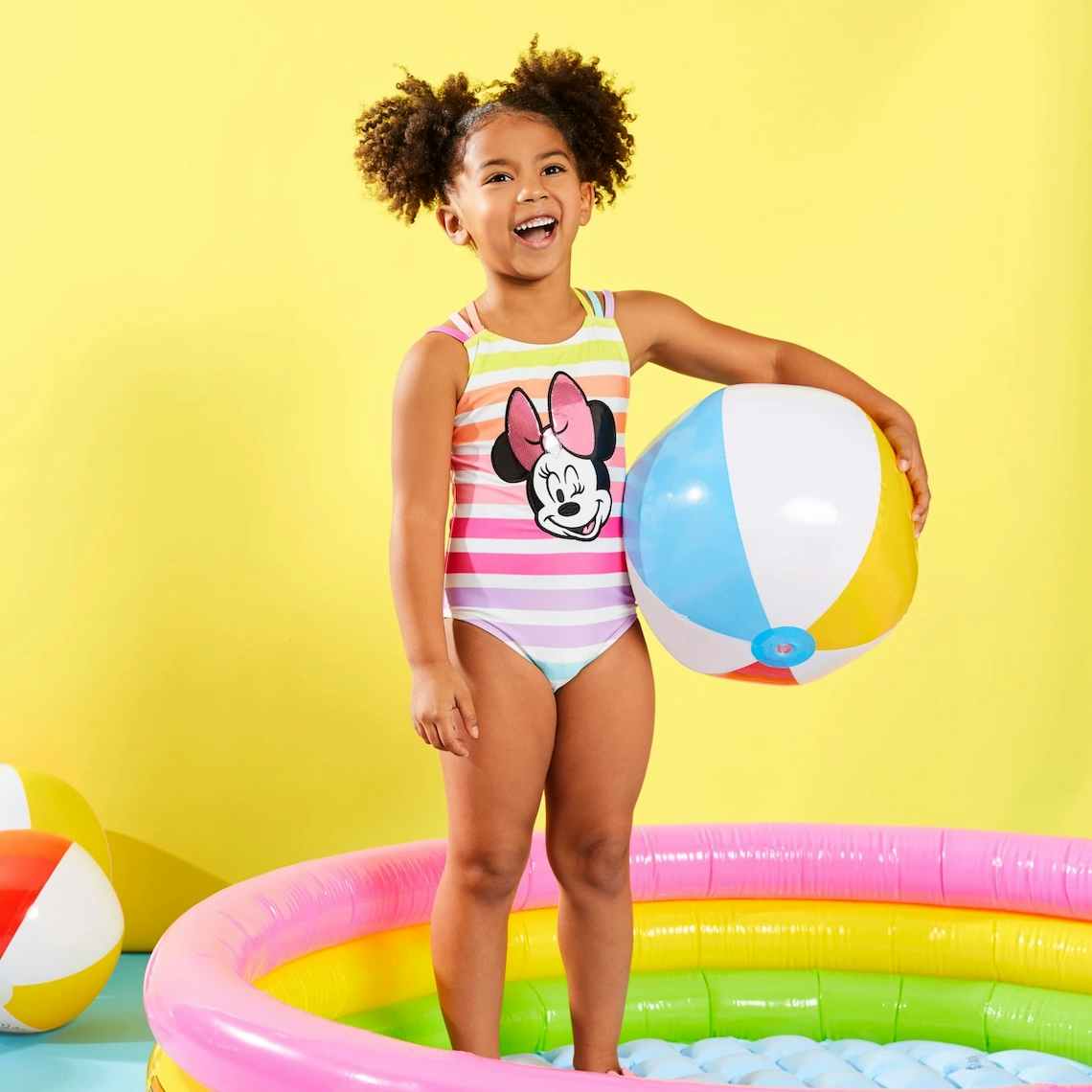 Little girl in swimsuit standing in kiddie pool holding a beach ball.
