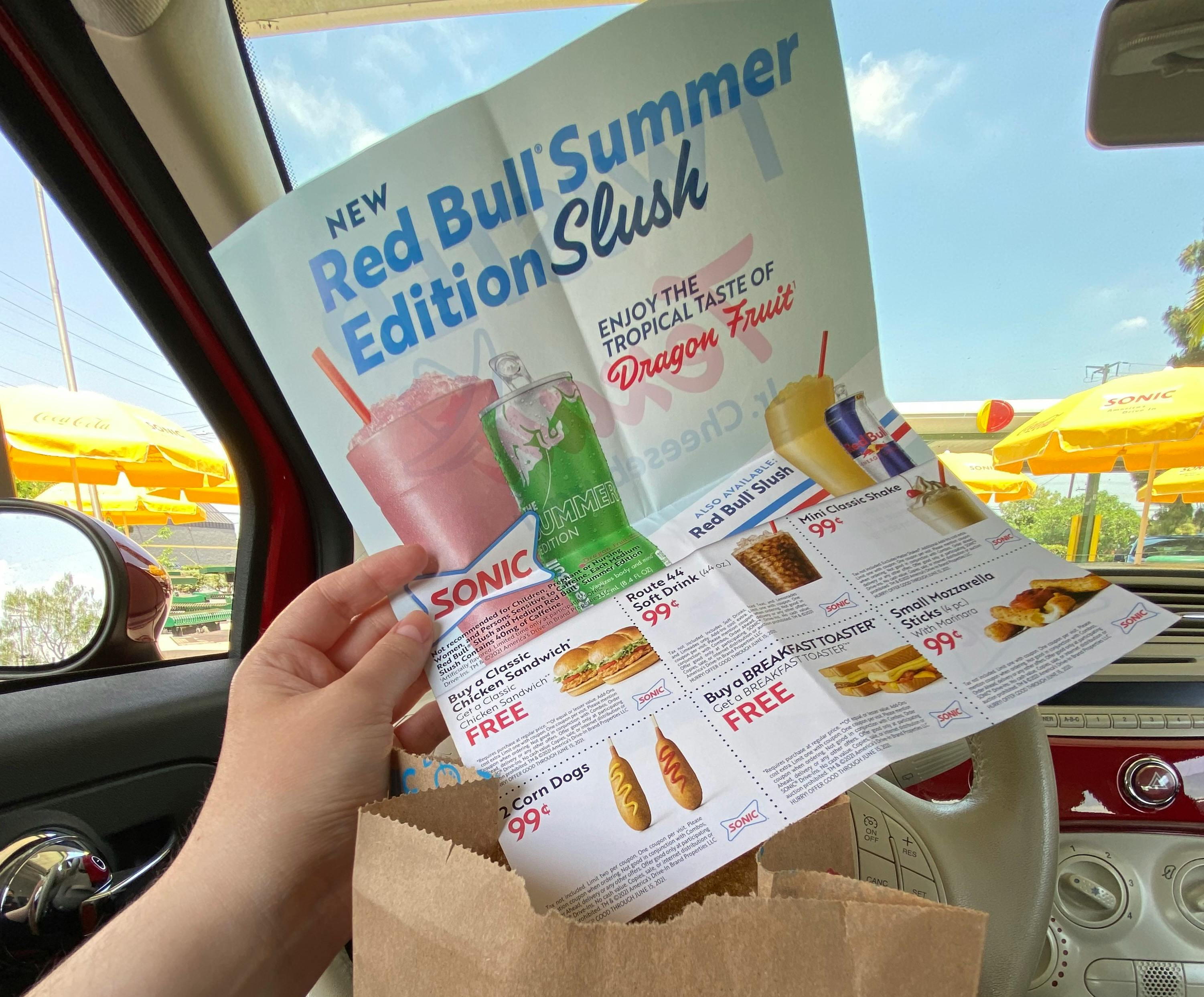 A person sitting in a car parked at Sonic, holding a page of Sonic coupons above a takeout bag.