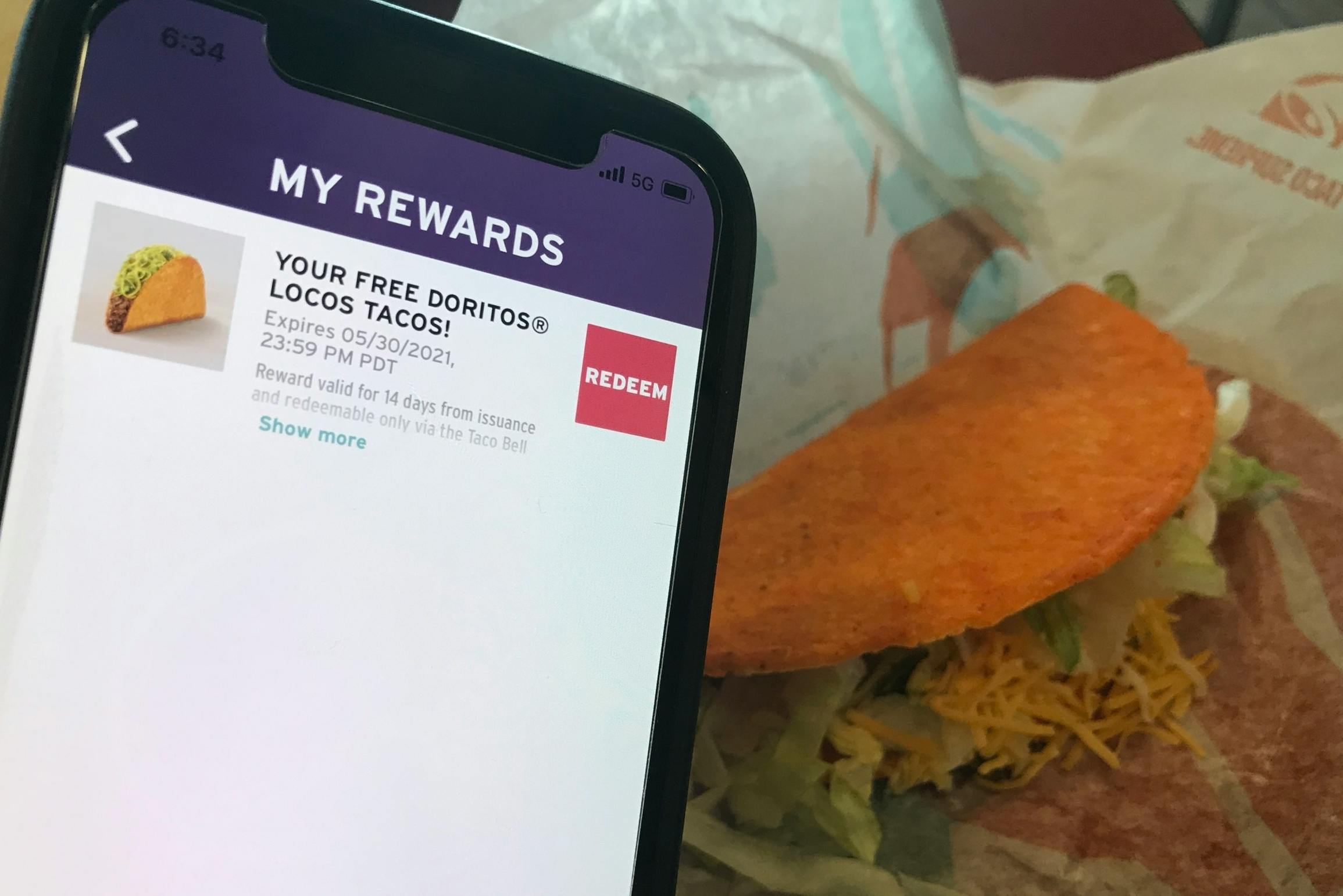 A cell phone displaying the Taco Bell app's My Rewards page, with a reward for a free Doritos Locos Tacos, being held in front of a Doritos Locos Tacos sitting on its paper wrapper.
