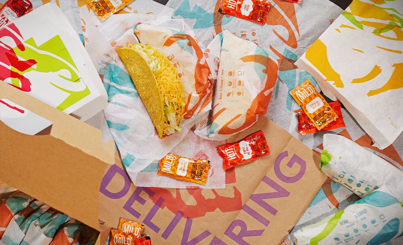 A pile of Taco Bell food and sauce packets with a Taco Bell bag that says "Delivering".