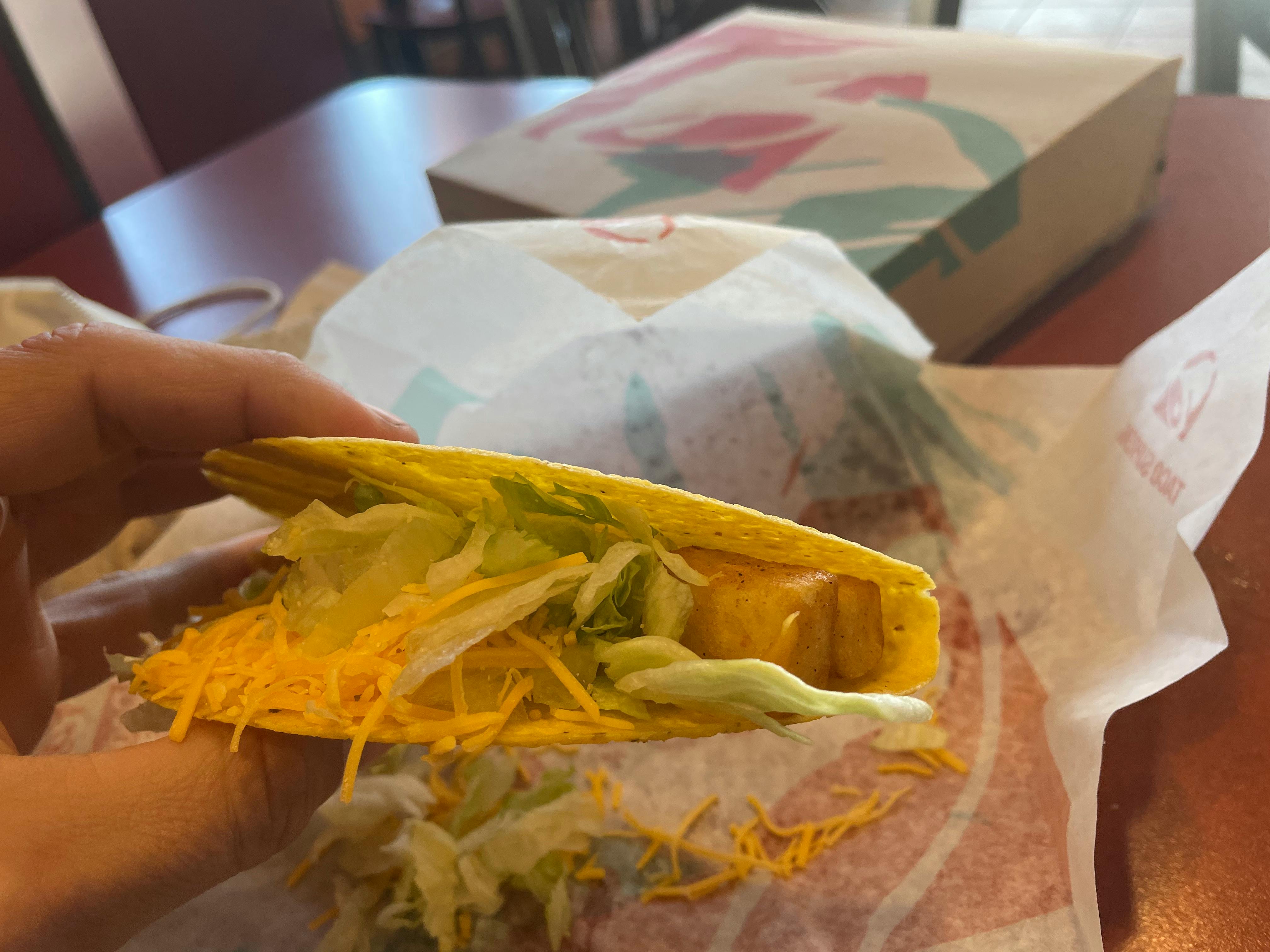 A person's hand holding a hard shell potato taco at a table inside Taco Bell.
