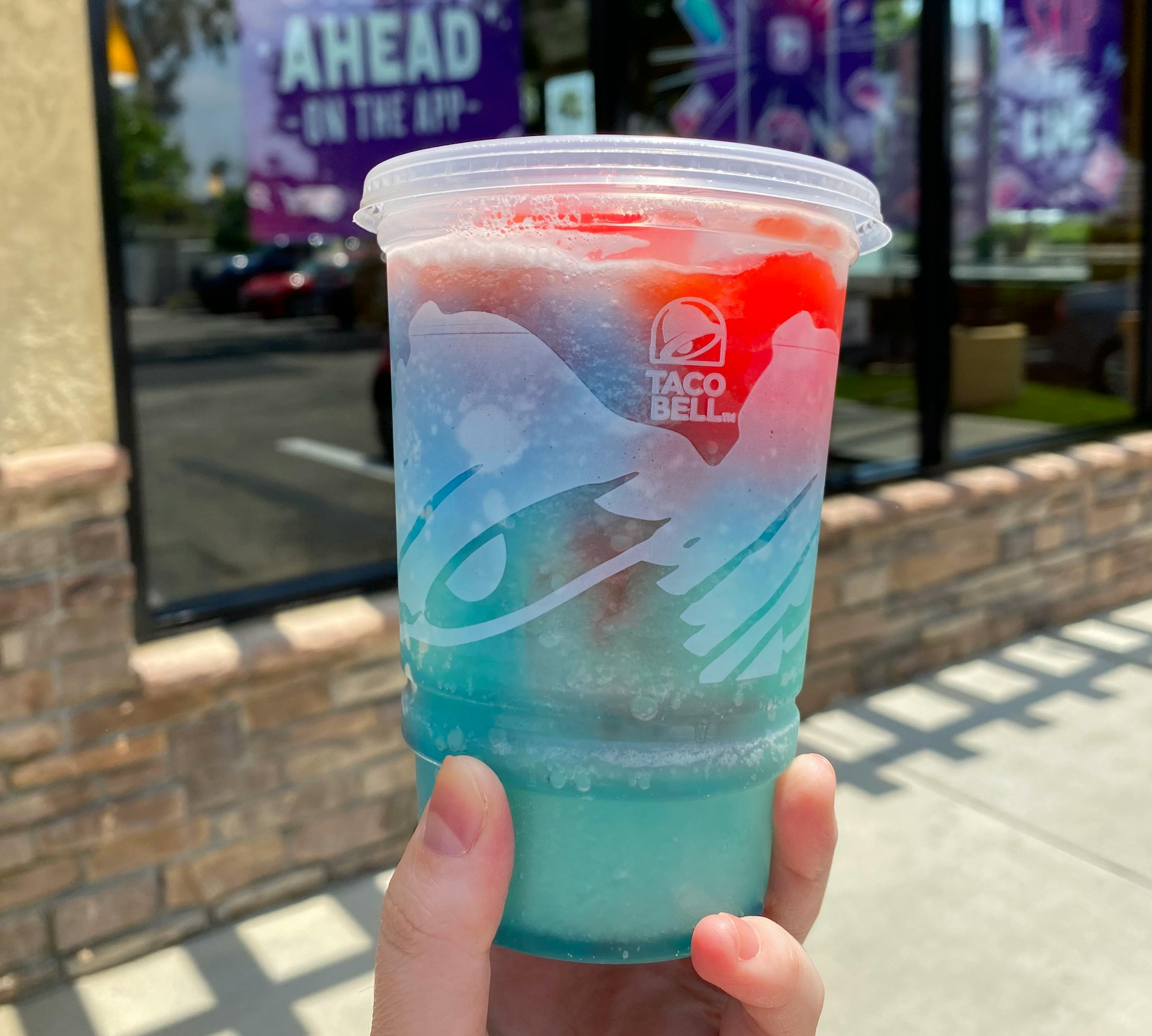 A person's hand holding a small Taco Bell slushie with layered flavors outside a Taco Bell restaurant.