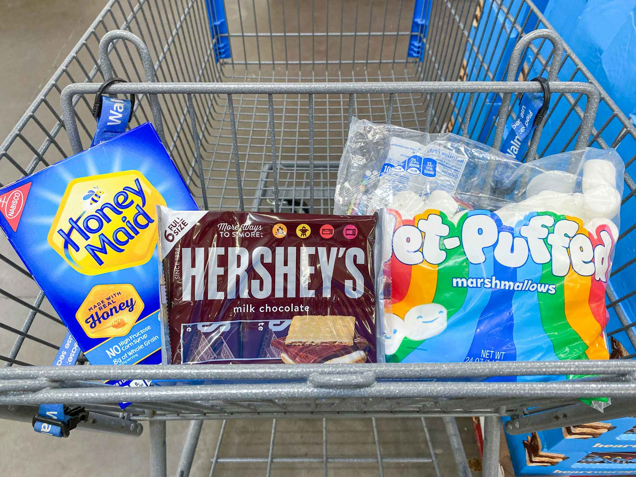 Walmart shopping cart with a box of honey maid graham crackers, six pack of full size hershey's chocolate bars, and a back of jet putffed marshmallows in the stop of the cart