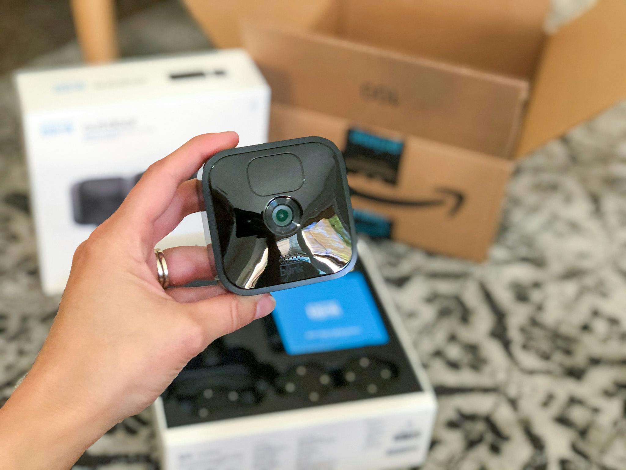 blink camera deals - A person's hand holding a Blink outdoor wireless security camera above its box next to an Amazon delivery box.