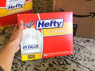 A hand holding a box of Hefty trash bags in front of an Amazon delivery box.