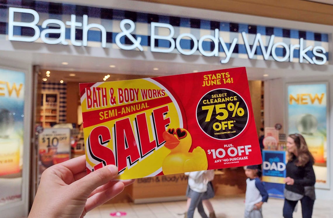 A person's hand holding up a Bath & Body Works Semi-Annual Sale coupon in front of a Bath & Body Works store entrance in a mall.