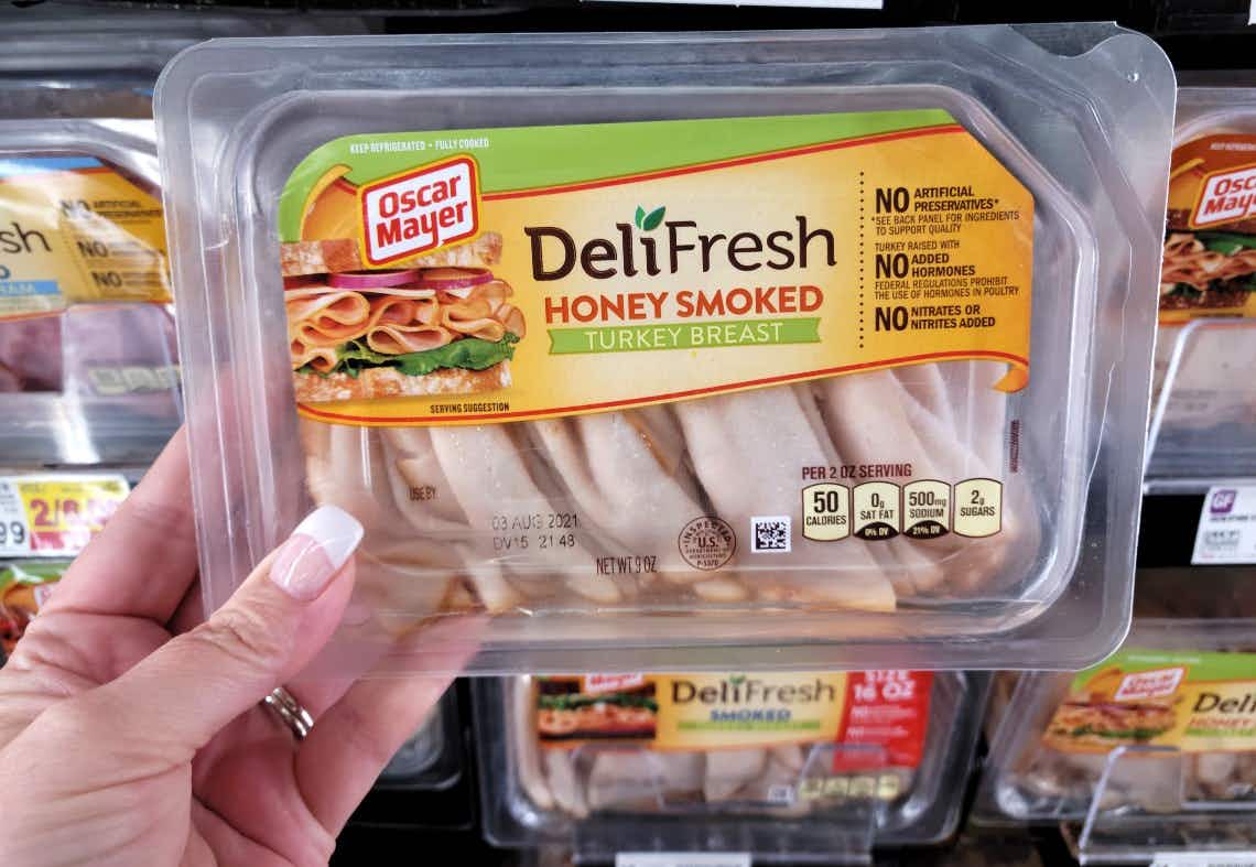 A package of Oscar Mayer DeliFresh honey smoked turkey breast being held in front of more packaged deli meat in a grocery store.