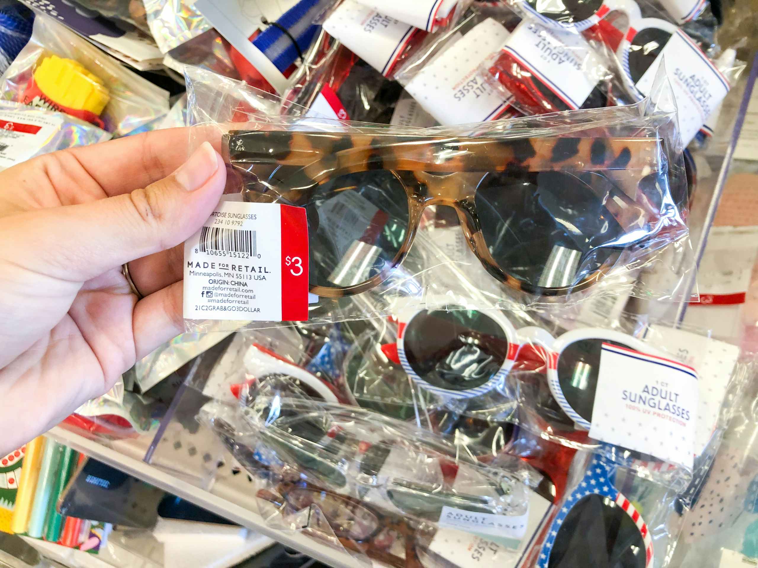 hand holding pair of plastic sunglasses over the top of a pile of sunglasses, all packaged